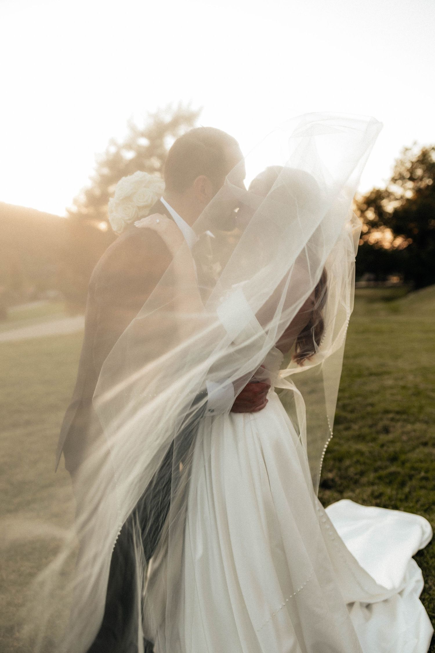 Romantic backlit photo of newlyweds kissing beneath a billowing veil in a sunlit outdoor setting.