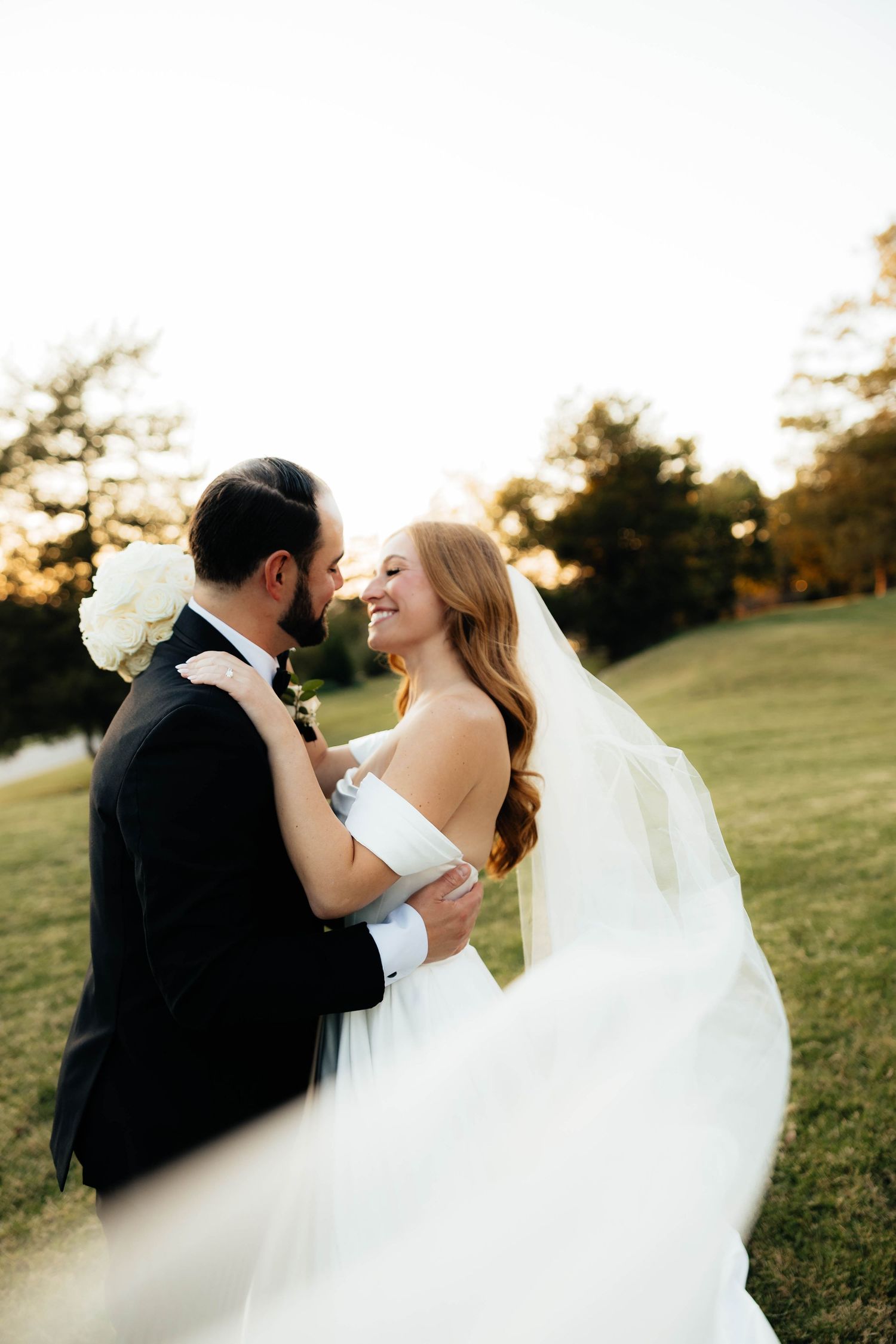 Bride and groom embracing at golden hour on a green lawn with veil flowing in wind.