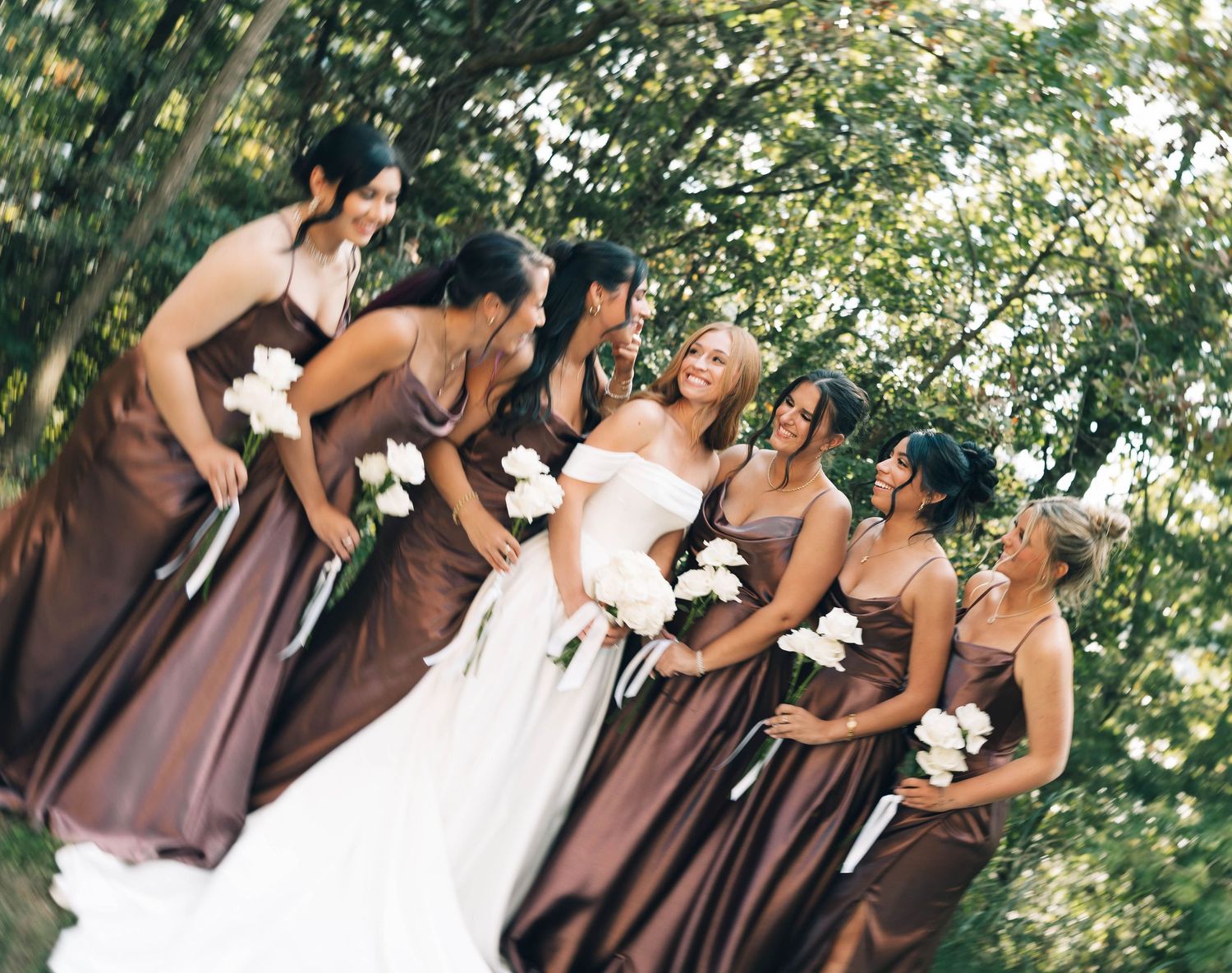 Bride in white off-shoulder gown laughs with bridesmaids in brown satin dresses under green trees.