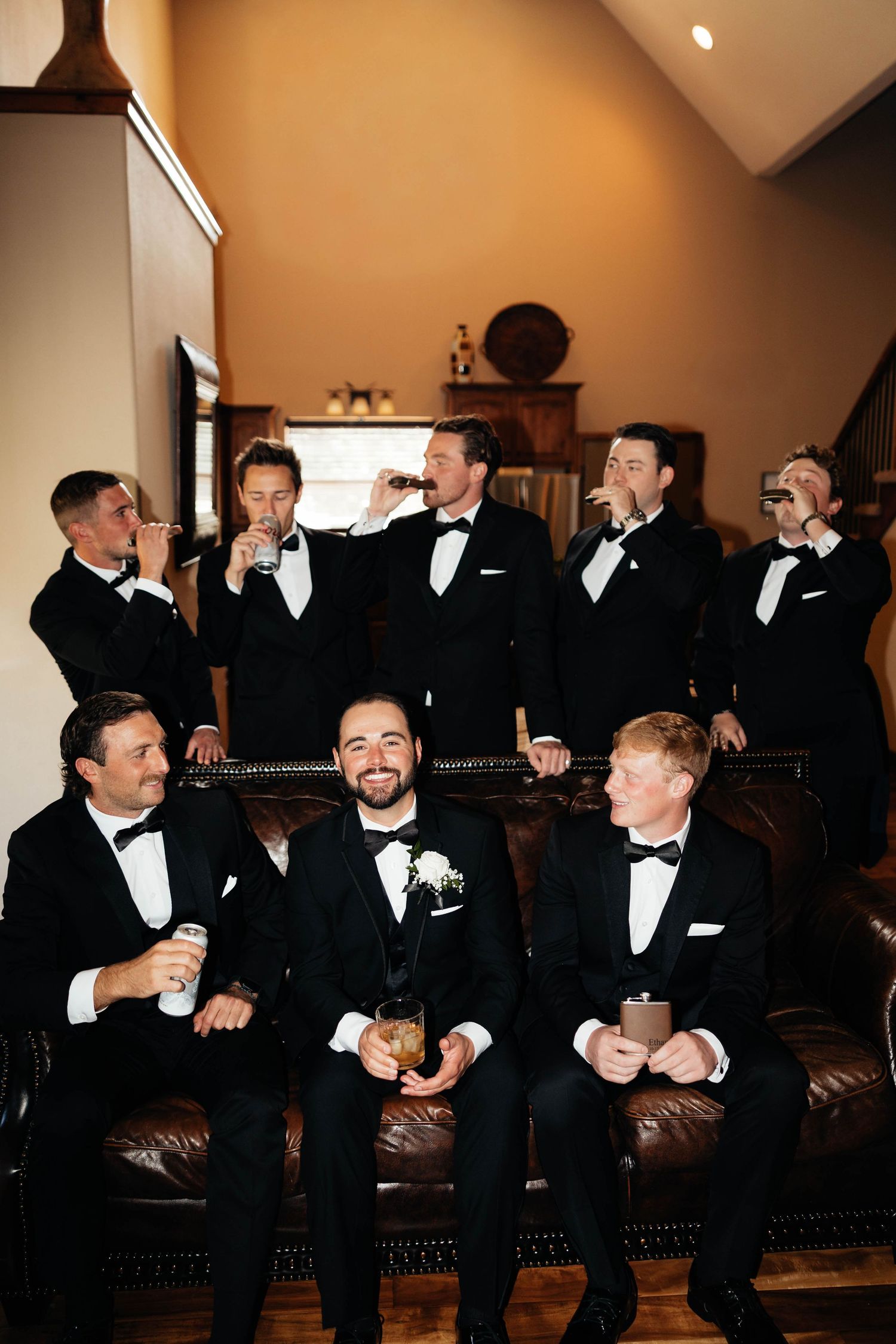 Groom and groomsmen in black tuxedos celebrate together in a warmly lit room.