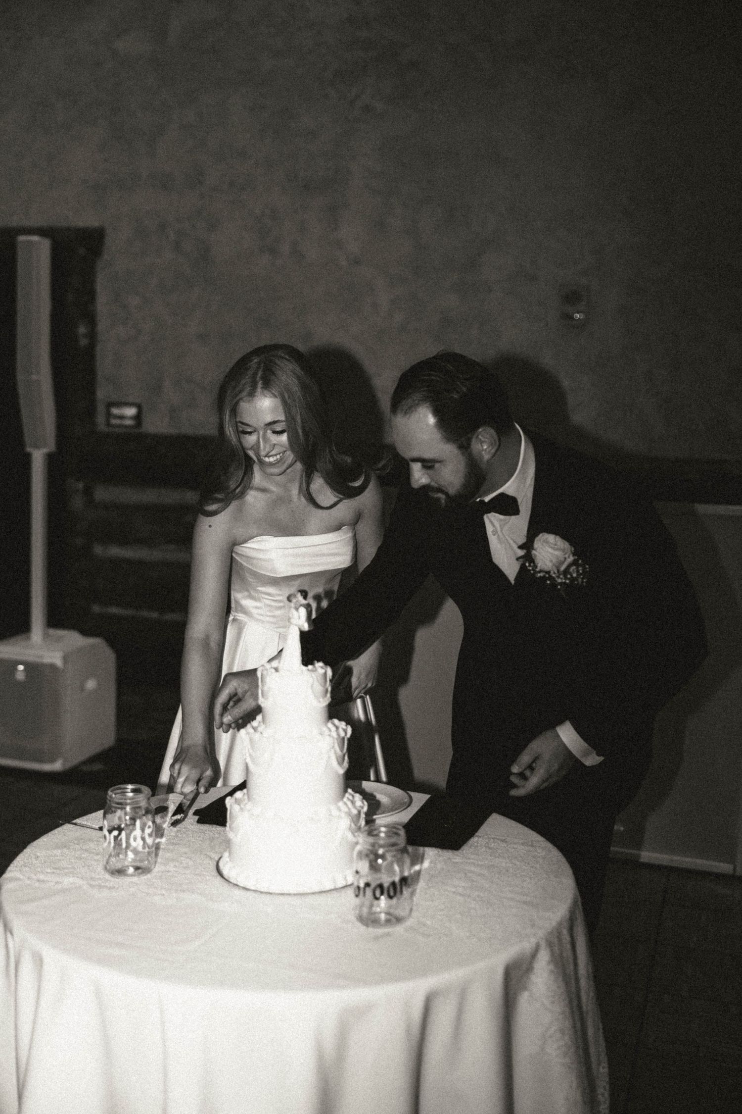 Black and white photo of smiling bride and groom cutting their three-tier white wedding cake together.