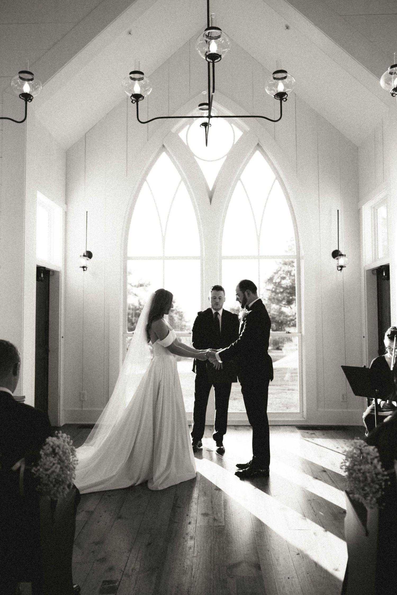 Black and white silhouette of bride and groom exchanging vows in stunning white chapel with arched windows.