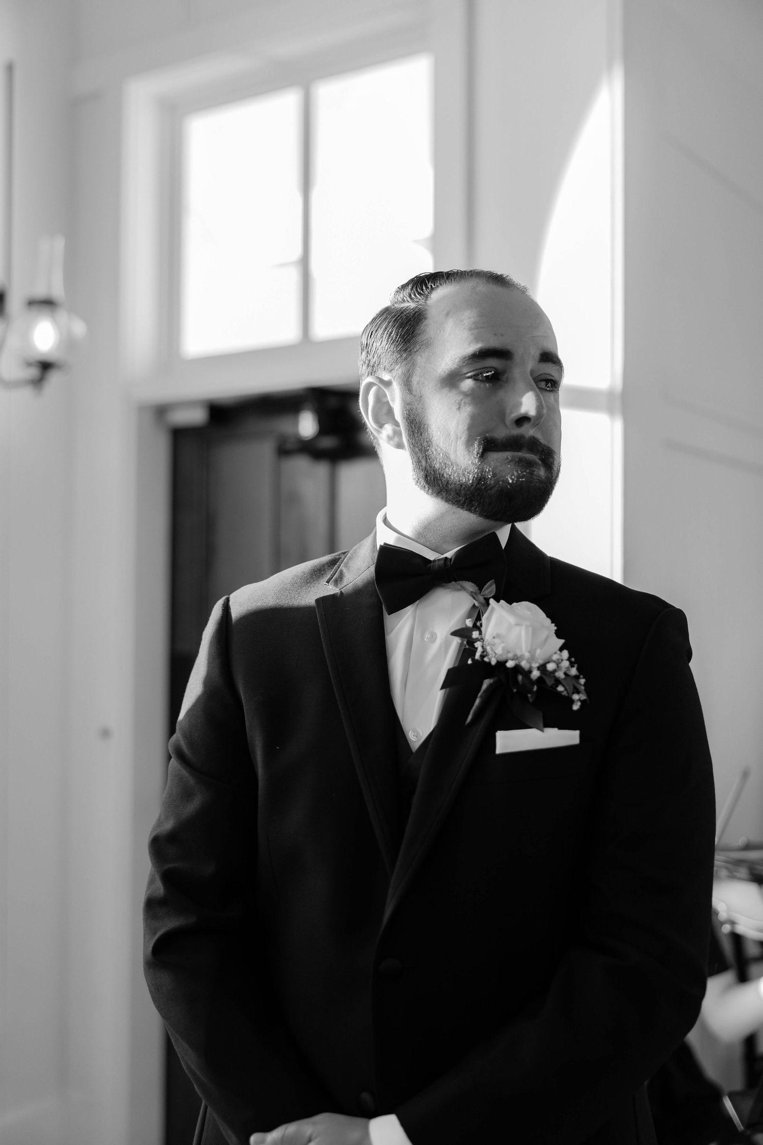 Groom in black tuxedo with boutonniere standing in church light, black and white photo.
