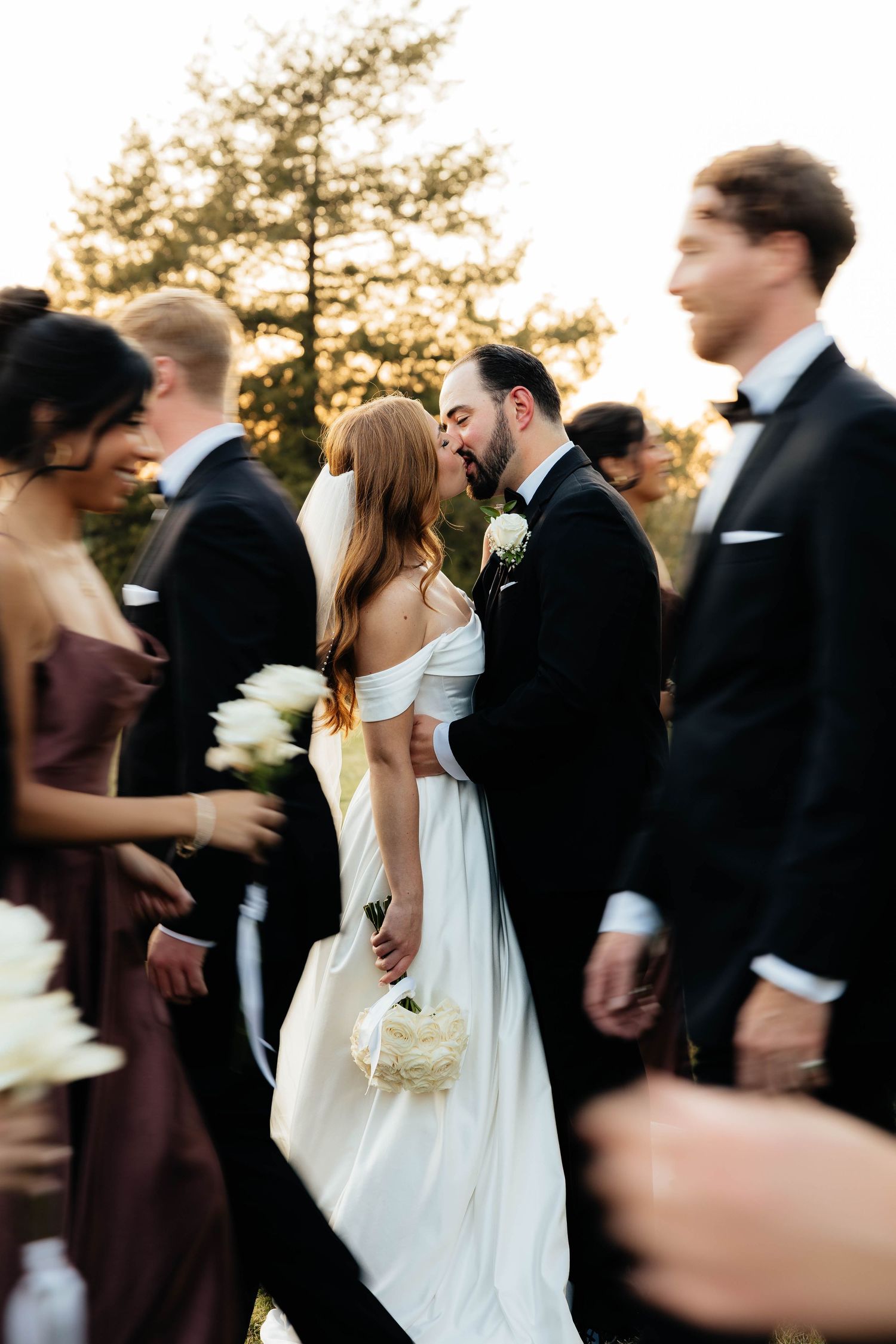 Bride and groom sharing a kiss surrounded by wedding party in elegant outdoor ceremony.