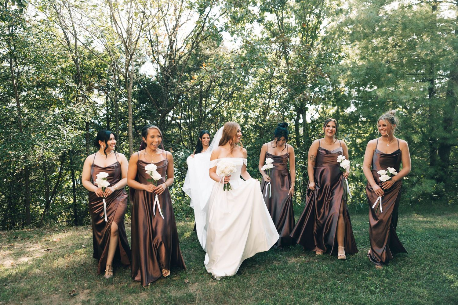 Bride in white gown walks with six bridesmaids in brown satin dresses through a green wooded area.