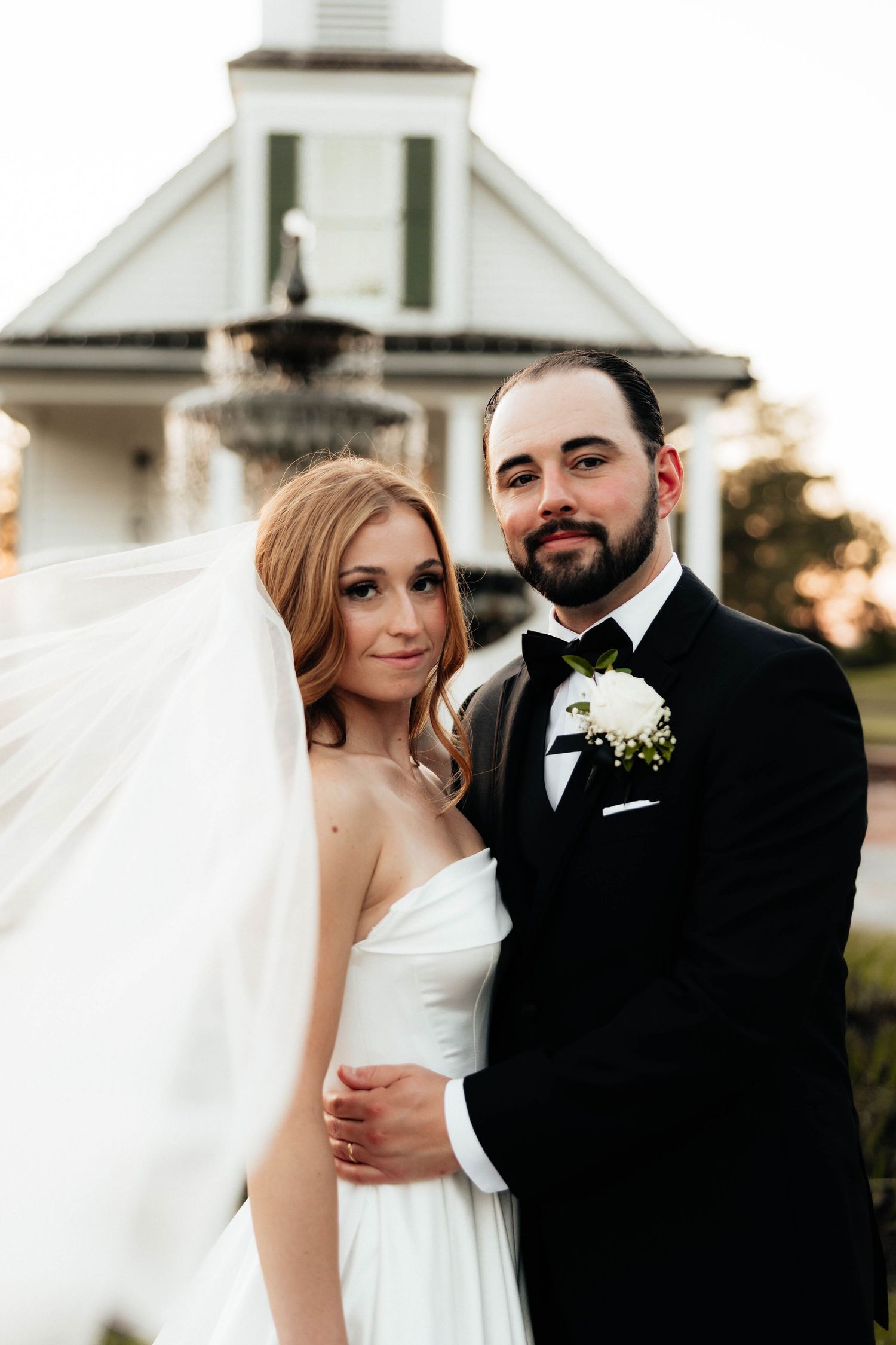 Elegant bride and bearded groom pose together near a white chapel fountain at golden hour.