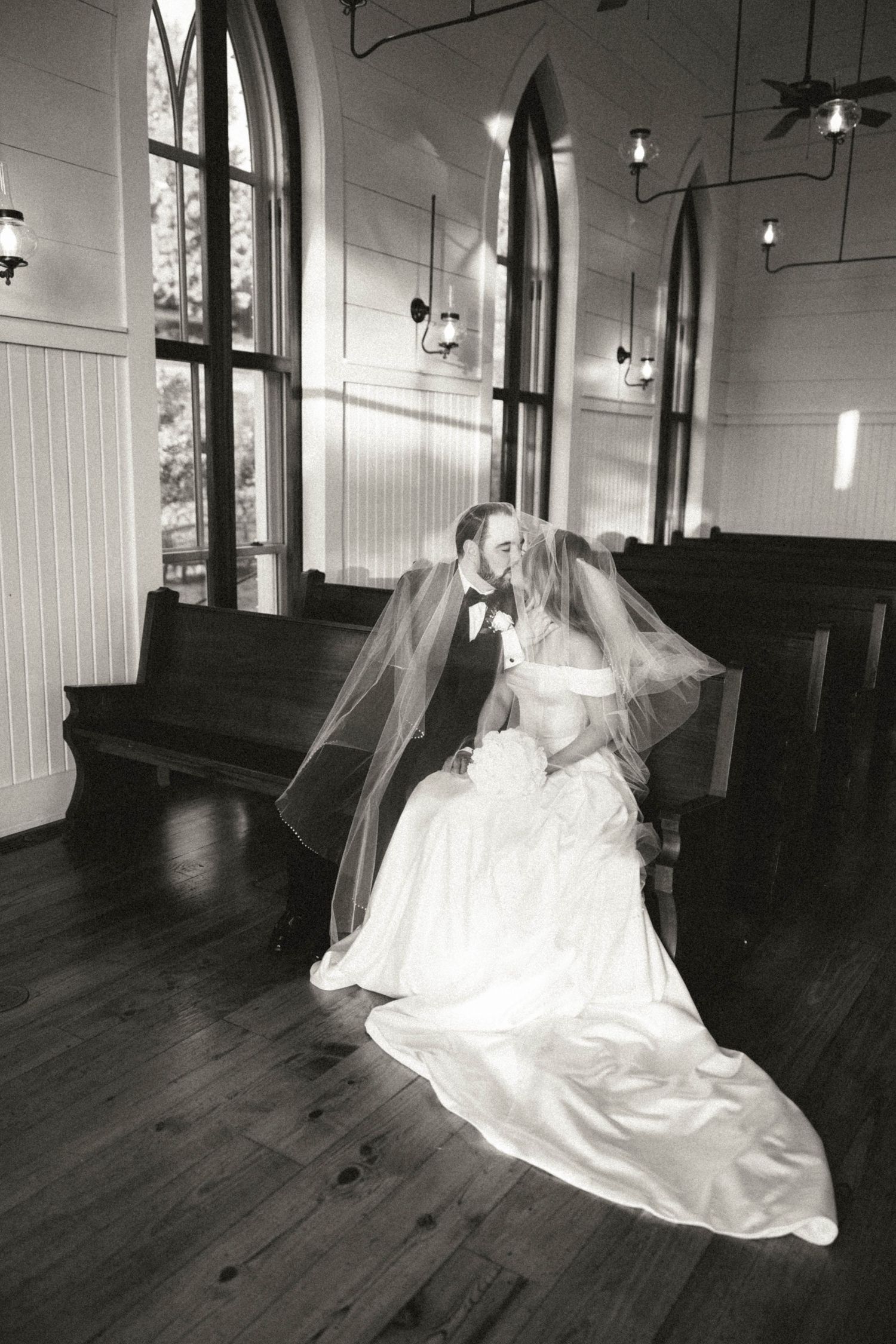Black and white photo of bride and groom sharing a kiss on a church pew beneath a flowing veil.