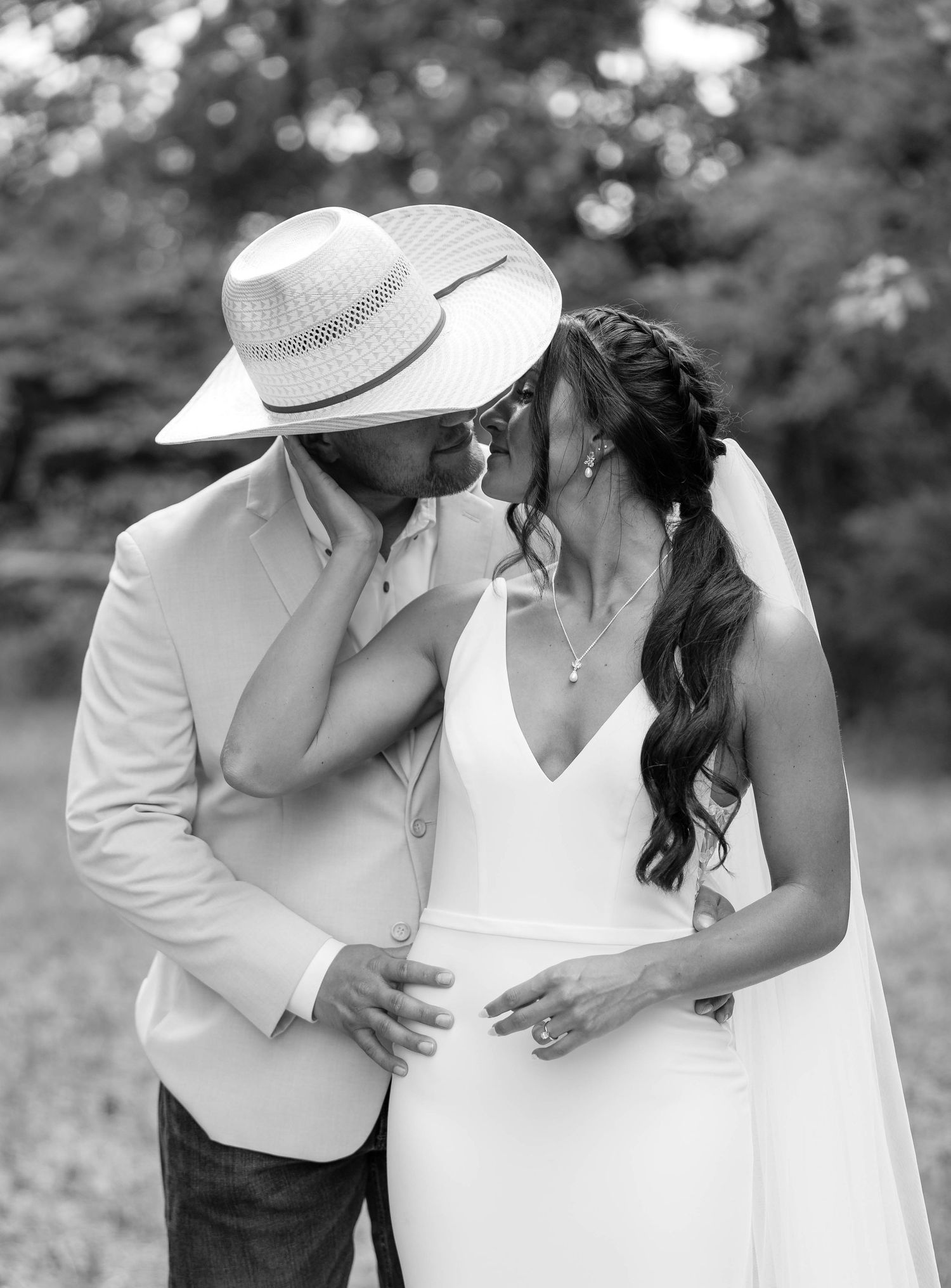 Black and white photo of groom in straw hat kissing bride in sleek white wedding dress outdoors.