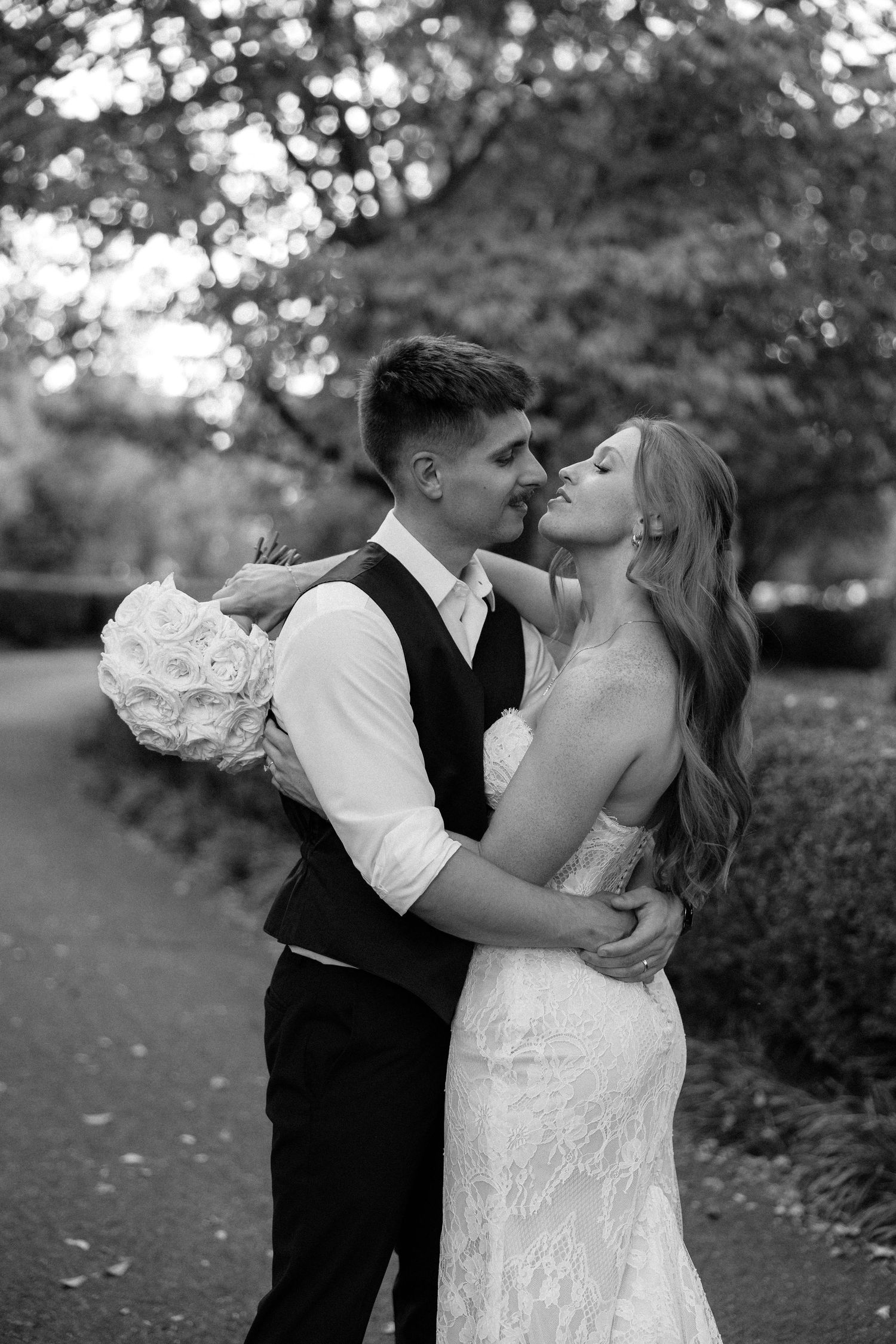 Black and white photo of bride and groom sharing a romantic kiss in a garden pathway.