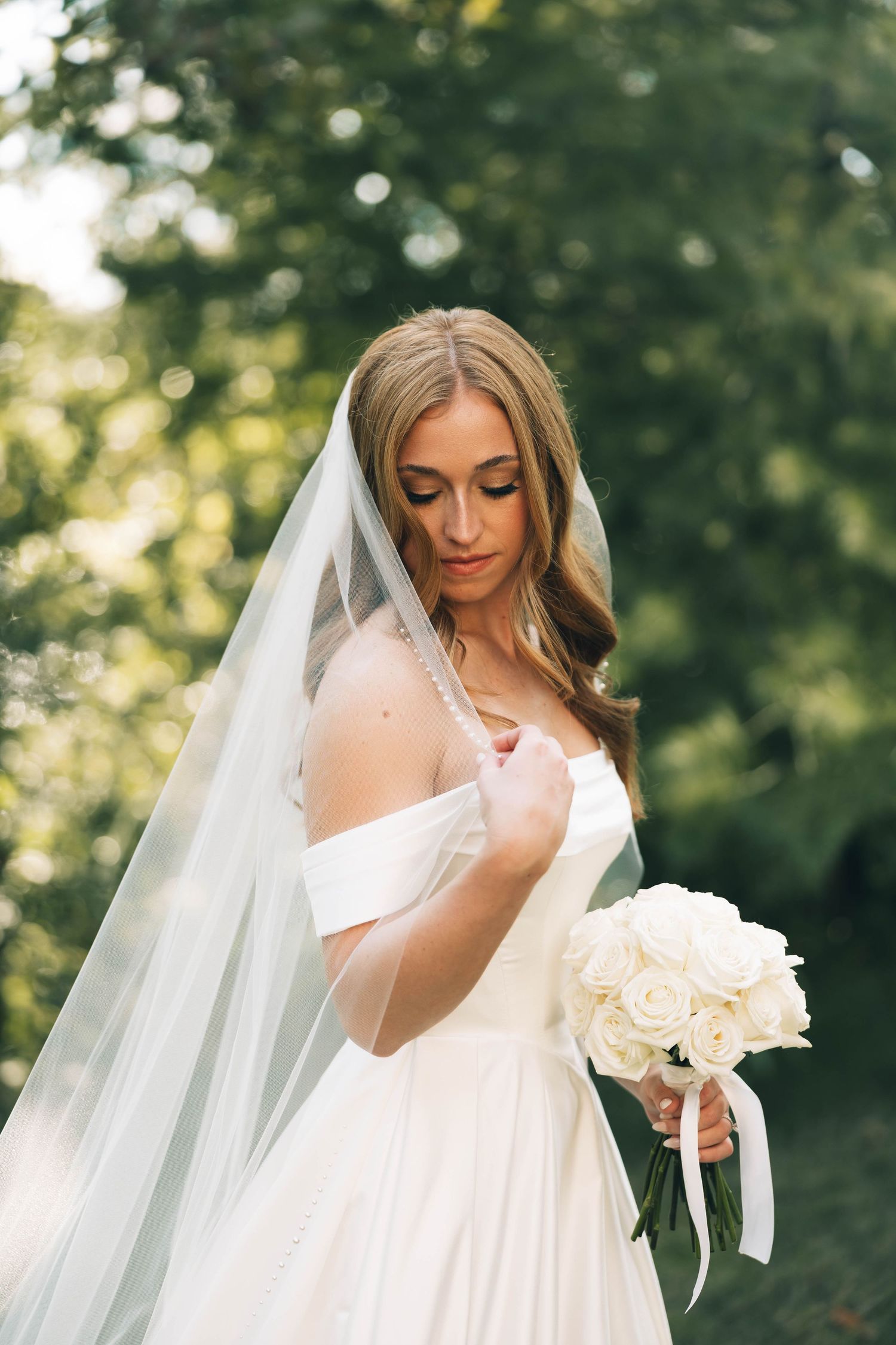 Beautiful bride in off-shoulder gown holding white rose bouquet under flowing veil outdoors.