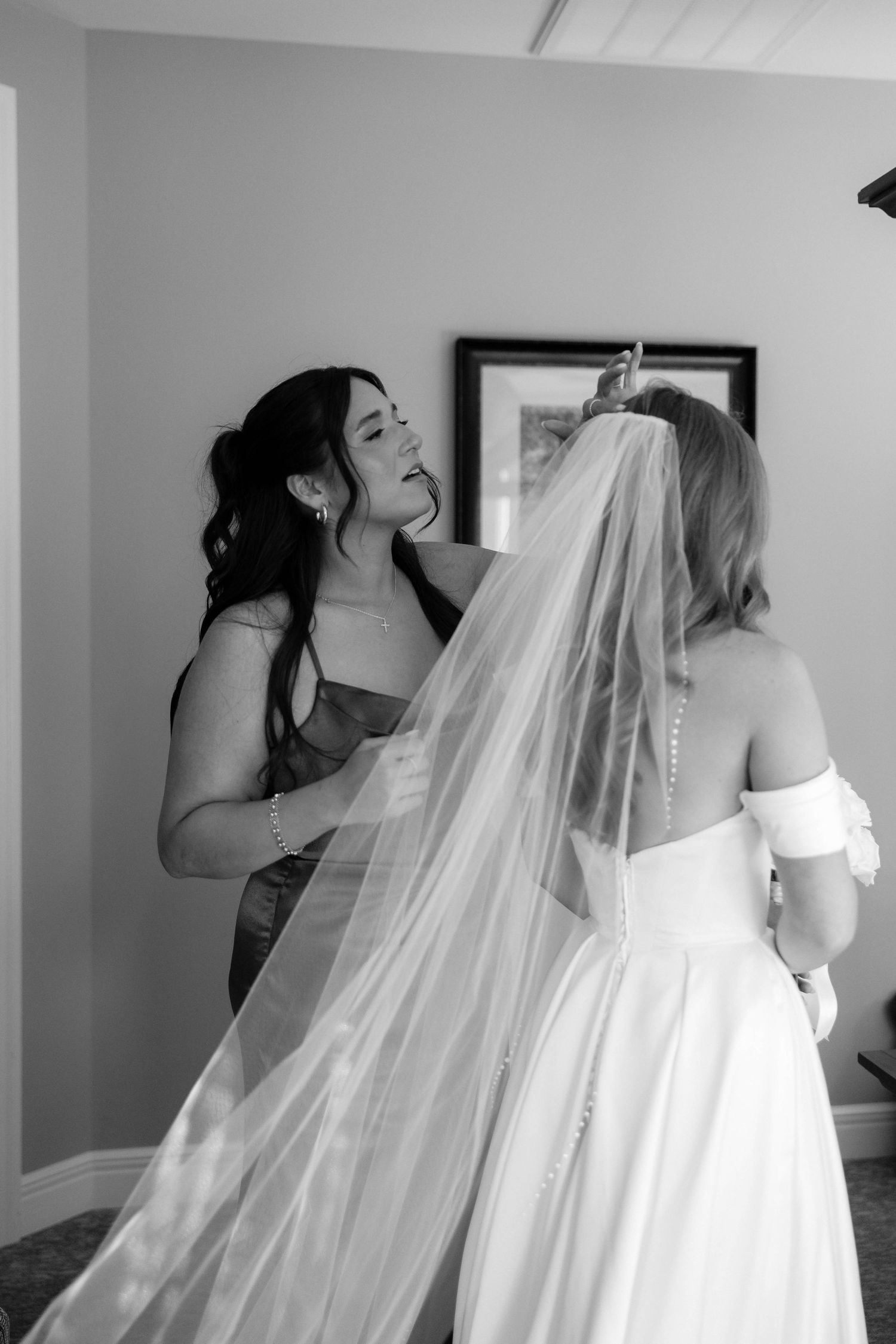 Black and white photo of bridesmaid emotionally helping bride adjust her cathedral veil before the wedding ceremony.