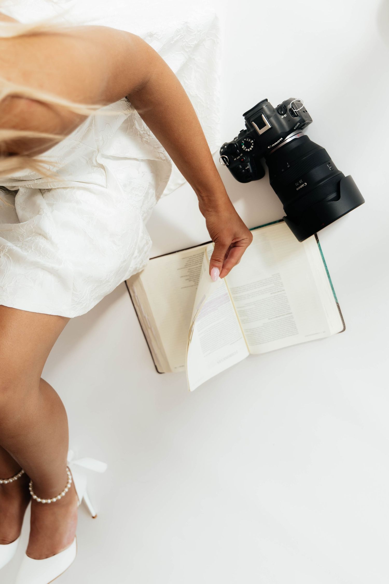 Overhead view of woman in white dress lying with open book and DSLR camera on white floor.