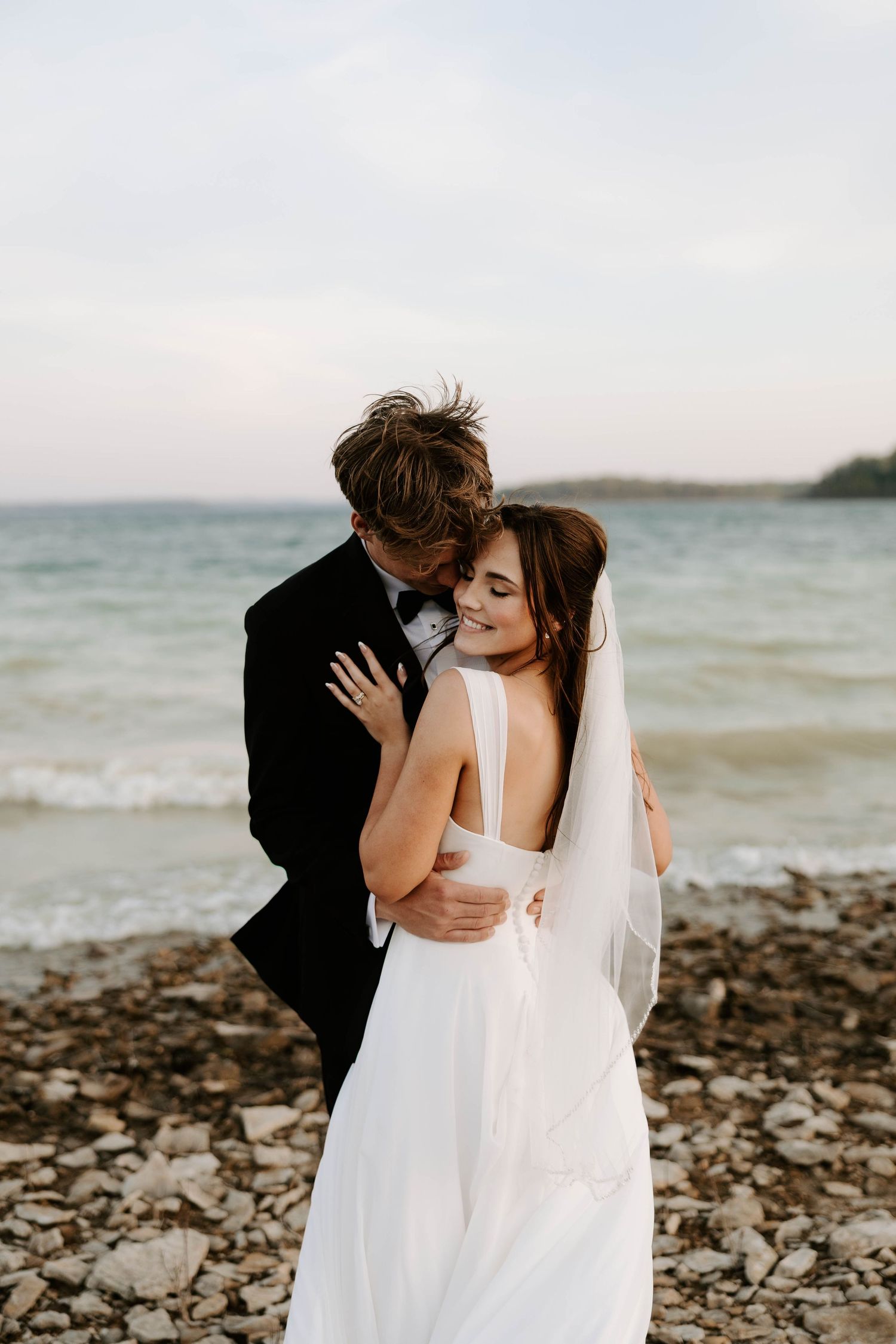 Bride and groom embracing on a rocky shoreline with calm ocean waters in the background.