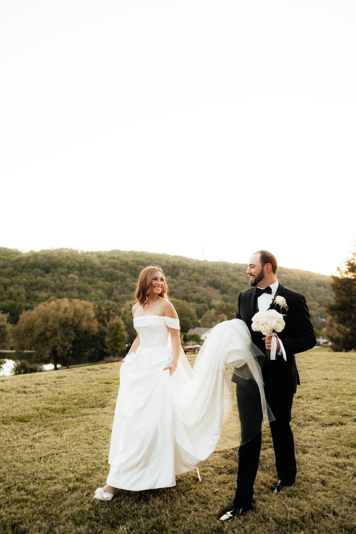 Bride and groom walk hand in hand on a hillside at dusk, groom carrying white bridal bouquet.