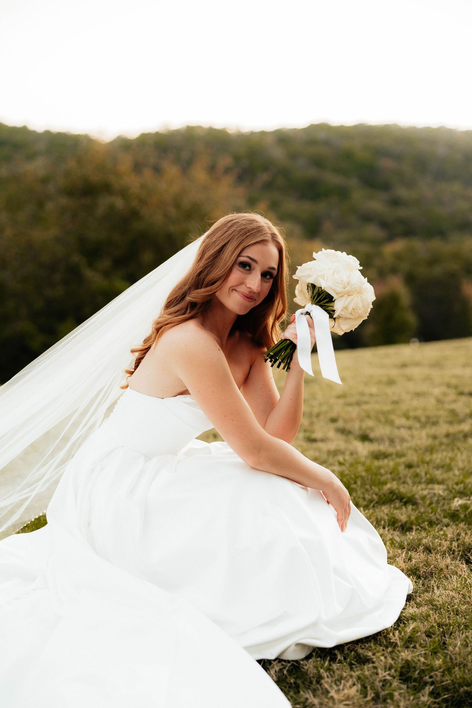 Smiling bride in strapless white gown with cathedral veil sitting on grass, holding white rose bouquet at golden hour.