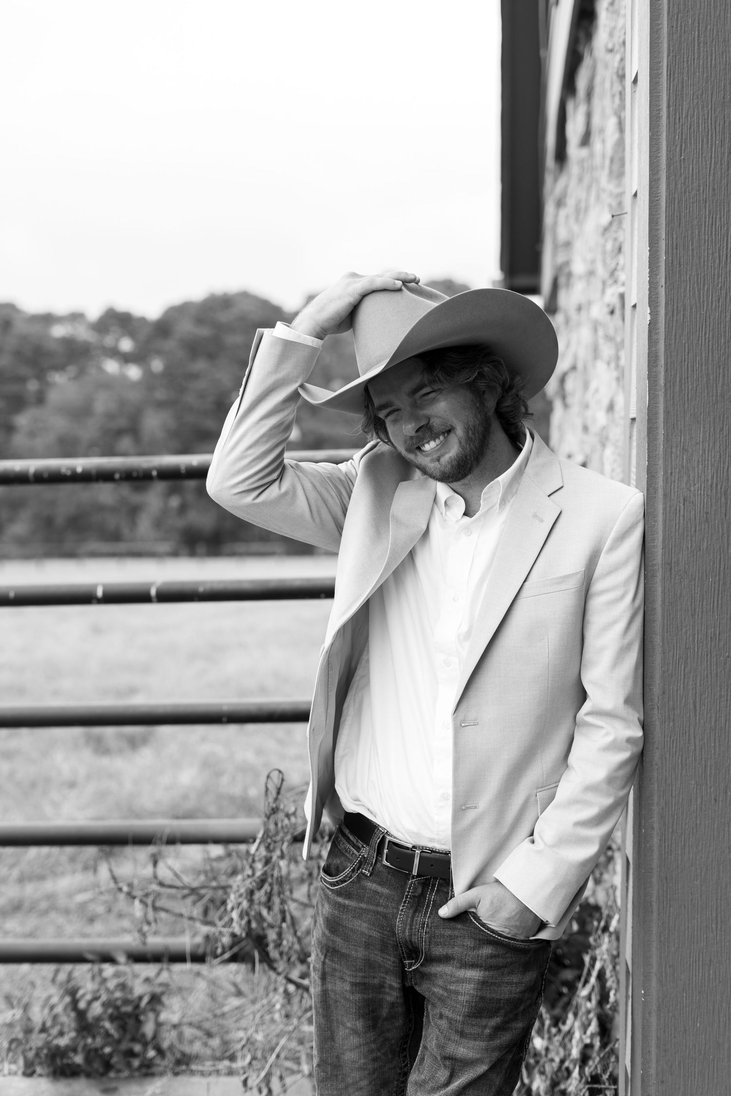 A person in a white blazer and cowboy hat poses against a rustic fence in black and white.