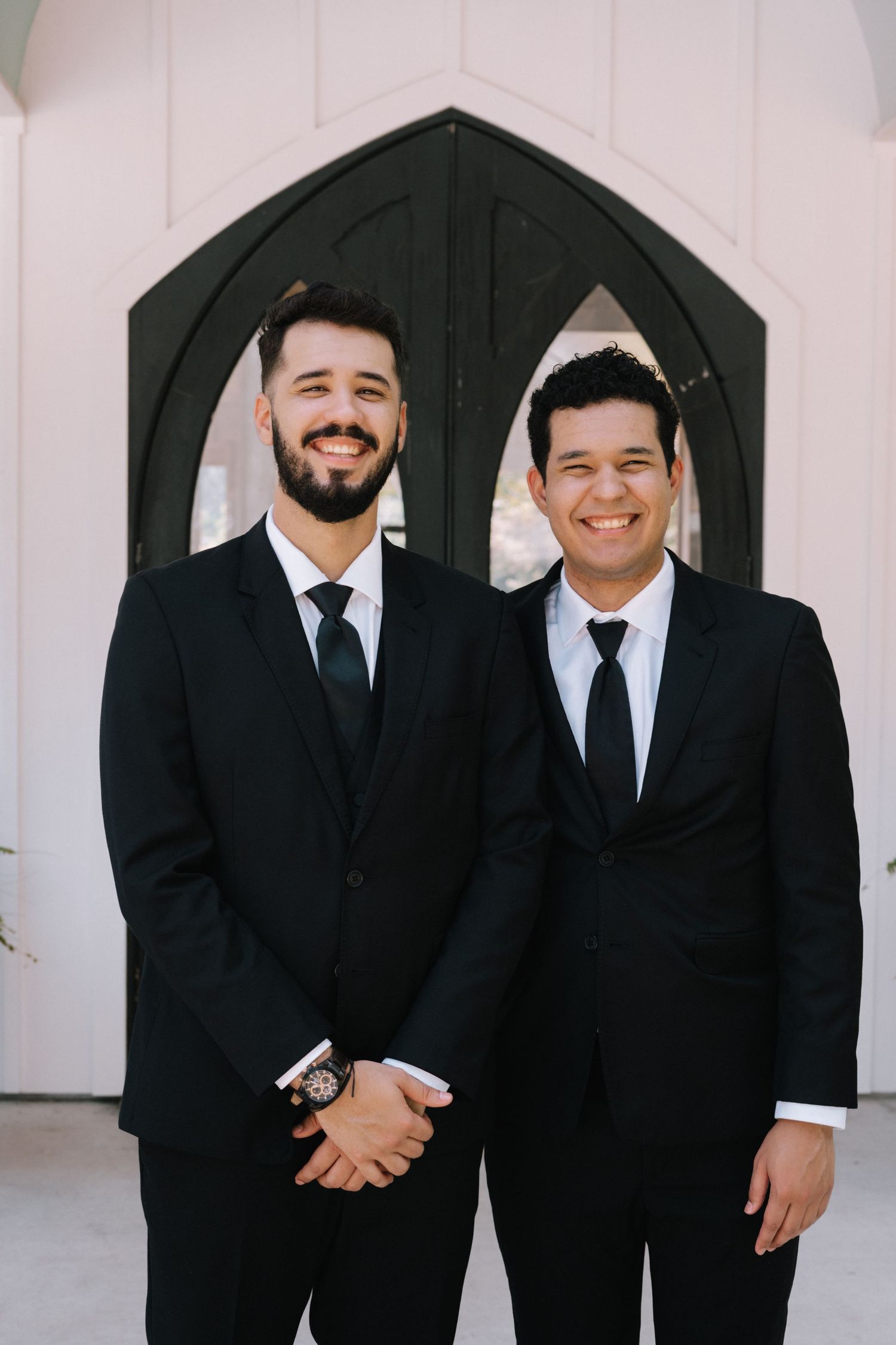 Groomsmen share laughs while posing in formal black suits outside a white chapel.