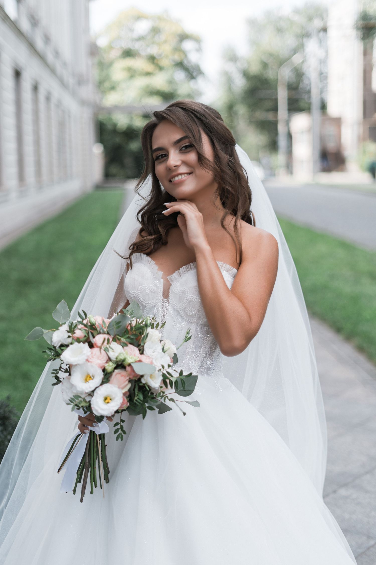 A bride in a white strapless gown and veil poses outdoors while holding a delicate bouquet of pink and white flowers.