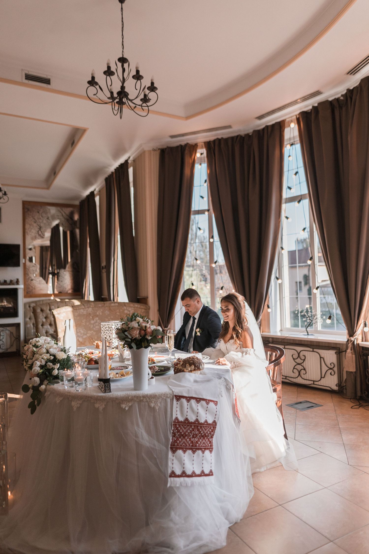 Elegant wedding reception table setting in a room with tall windows, chandelier and flowing brown curtains.