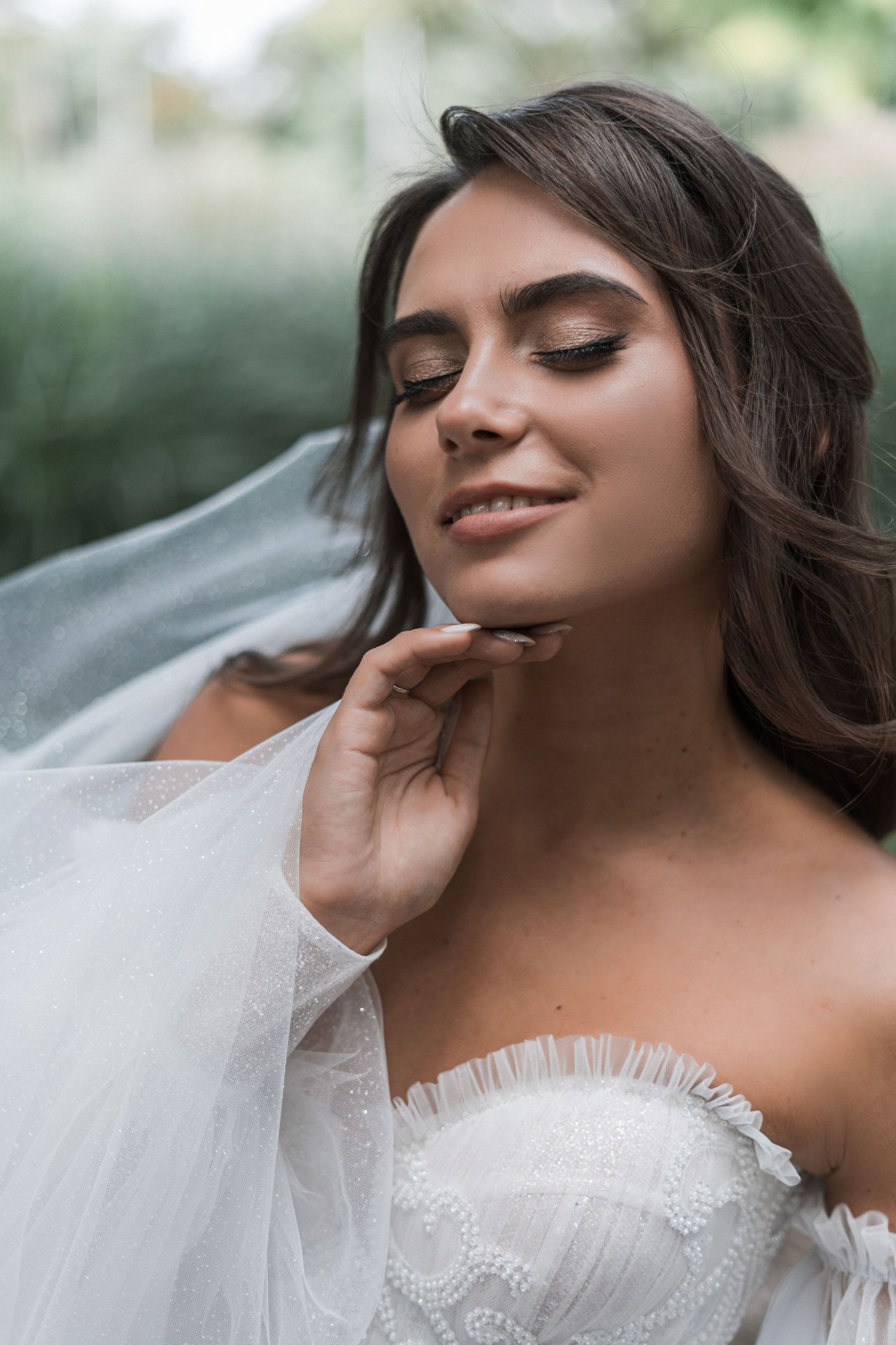 A bride in a white strapless wedding dress with delicate lace details poses with a flowing veil in an outdoor setting.