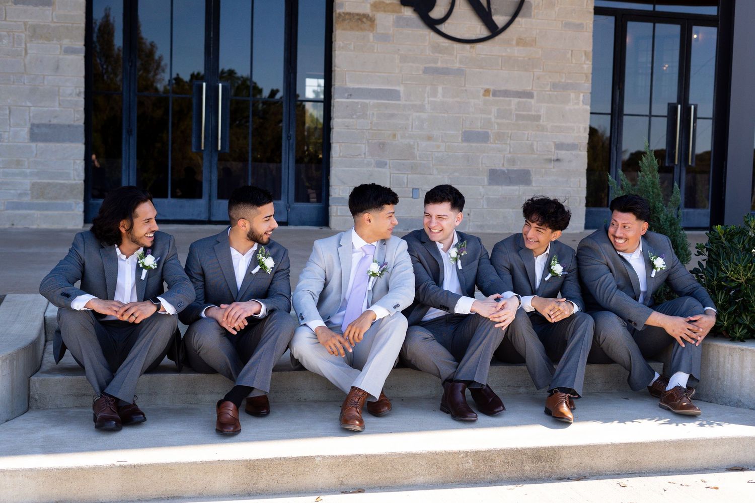 Group of groomsmen in gray suits sitting casually on stone steps outside.