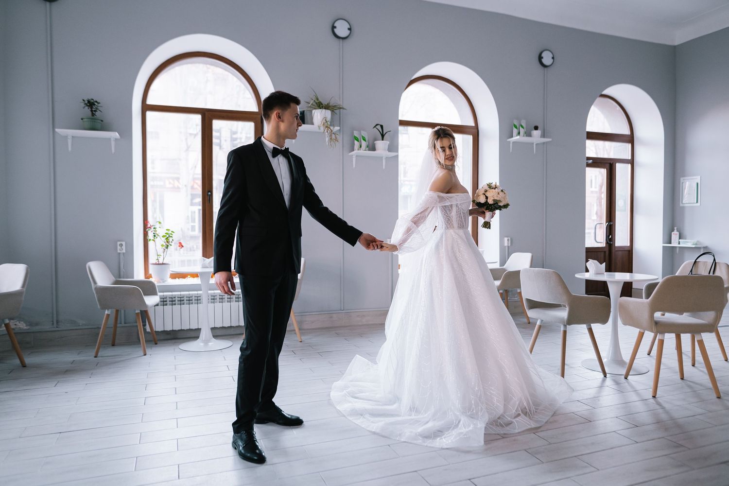 A couple in formal wedding attire dance together in an elegant room with arched windows and modern decor.