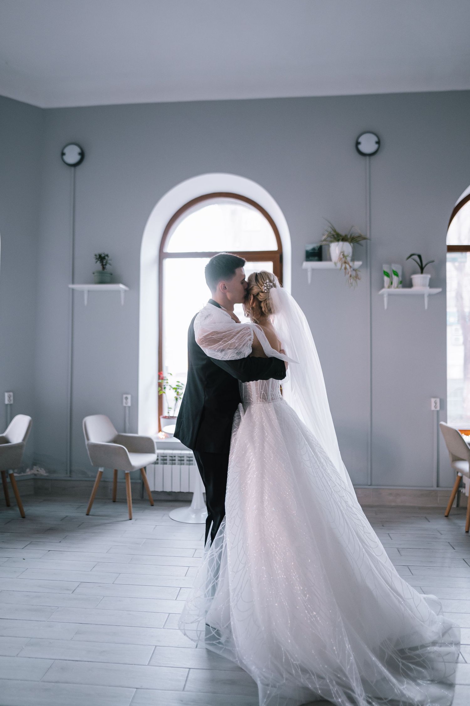 A romantic wedding moment captured in an elegant room with arched windows and white decor.