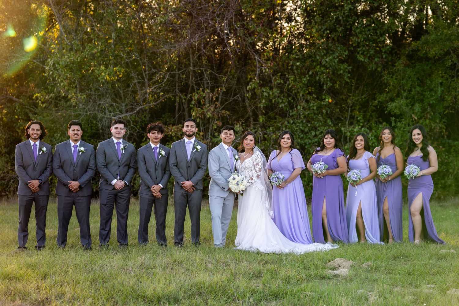 Wedding party in purple and gray attire poses together in a line outdoors.