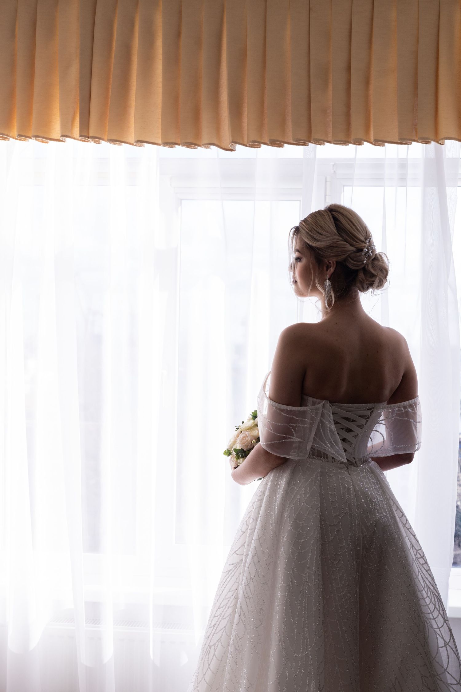 Elegant bride in off-shoulder white lace gown stands silhouetted against sheer curtains holding a small bouquet.
