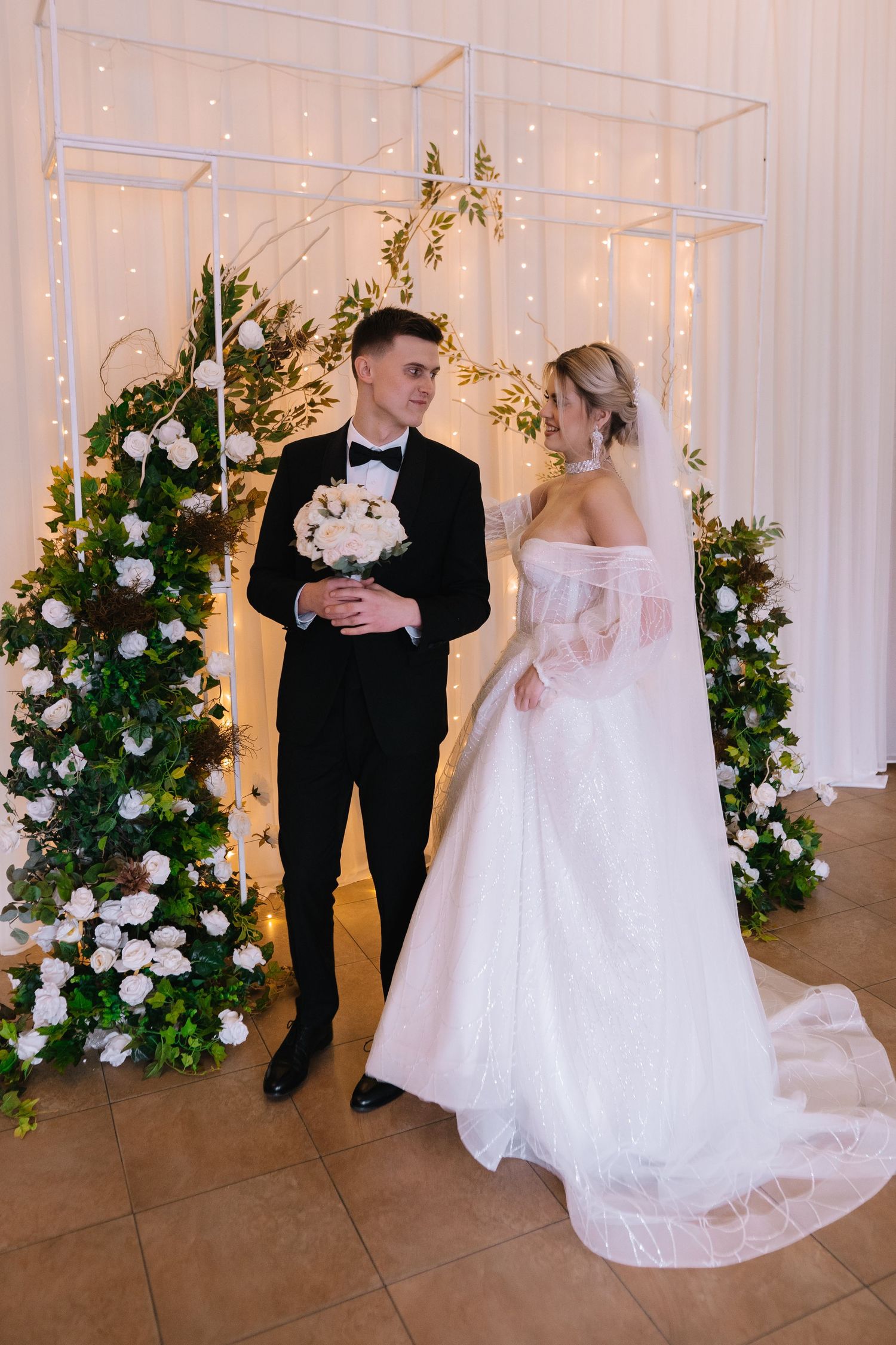 A couple shares their first dance at an elegant indoor wedding reception decorated with white floral arrangements and string lights.