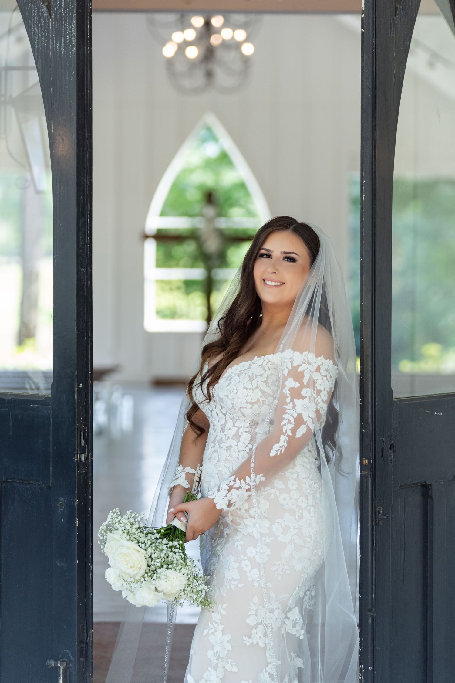 A bride in an elegant lace wedding dress with sheer details stands in front of dark French doors.