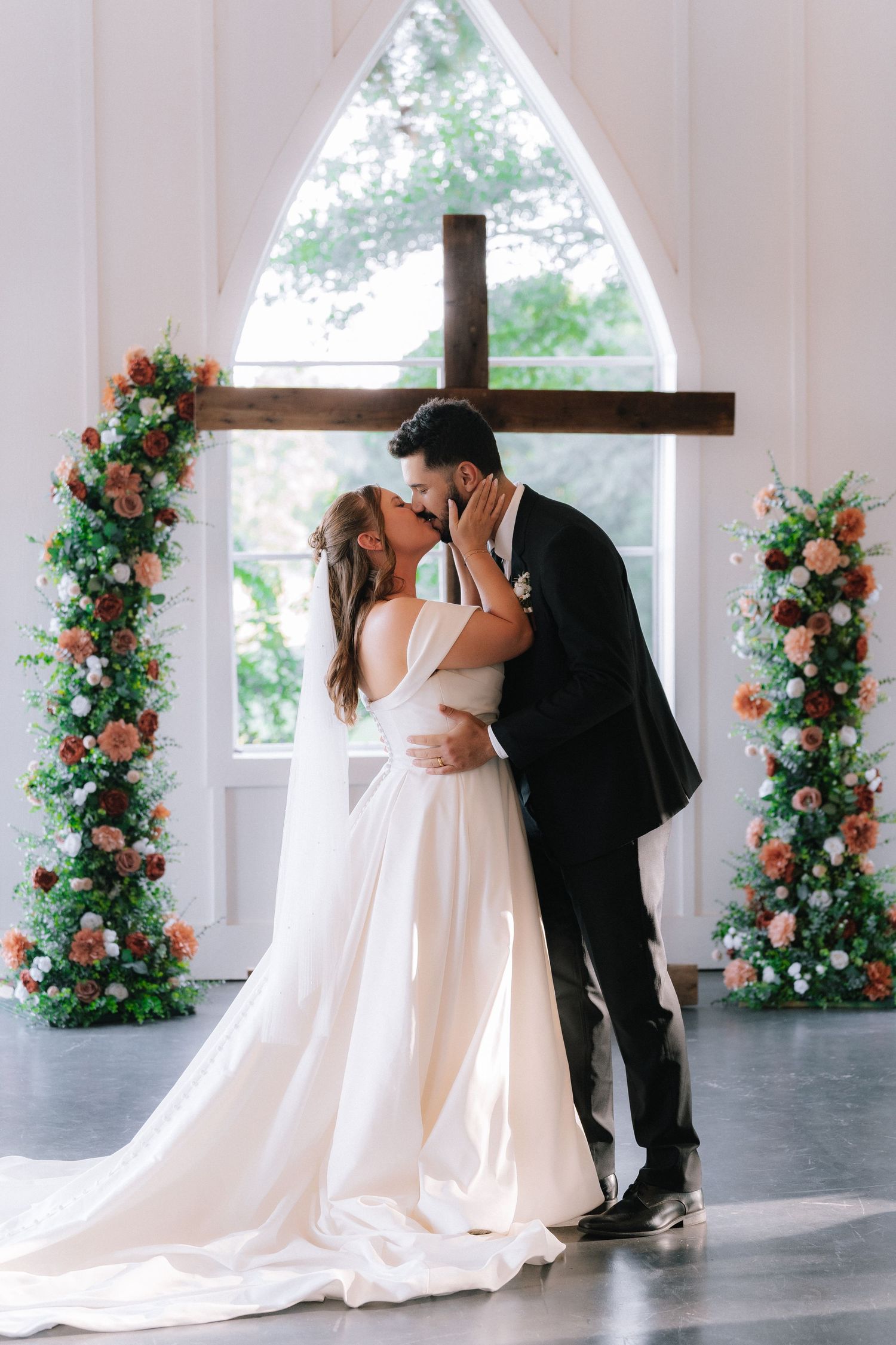 Wedding ceremony setup with floral arches and cross in a modern white chapel interior.