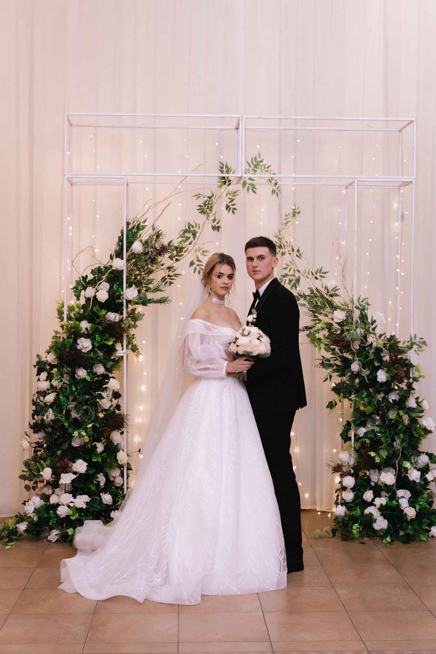 Elegant wedding arch decorated with white roses and greenery features a couple posing in formal attire against a light backdrop.