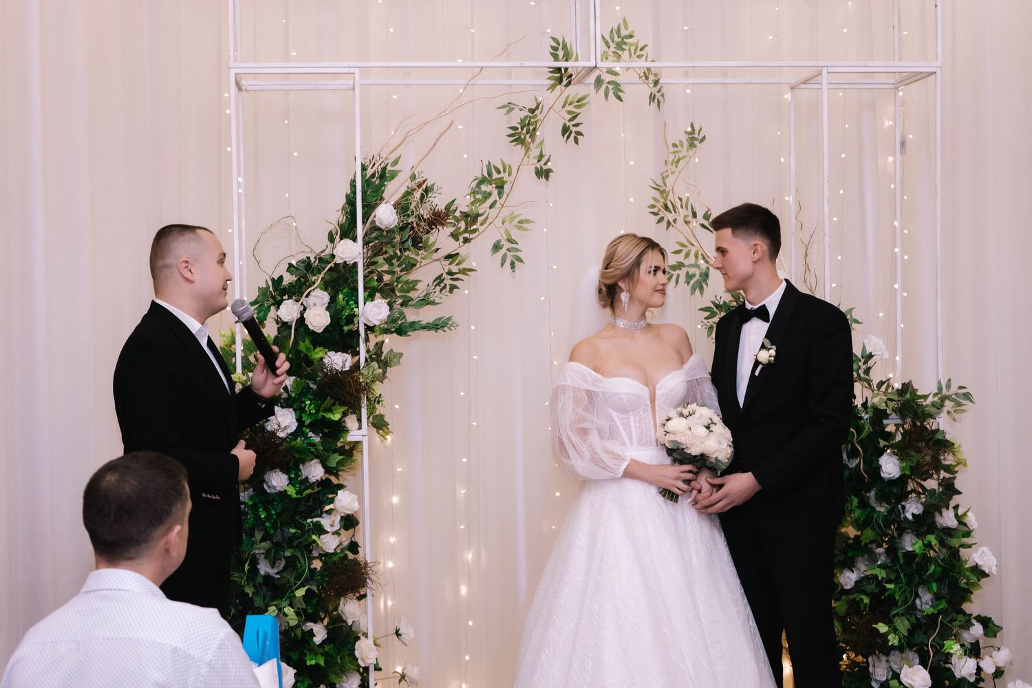 A wedding ceremony with floral decorations and string lights against a white backdrop.