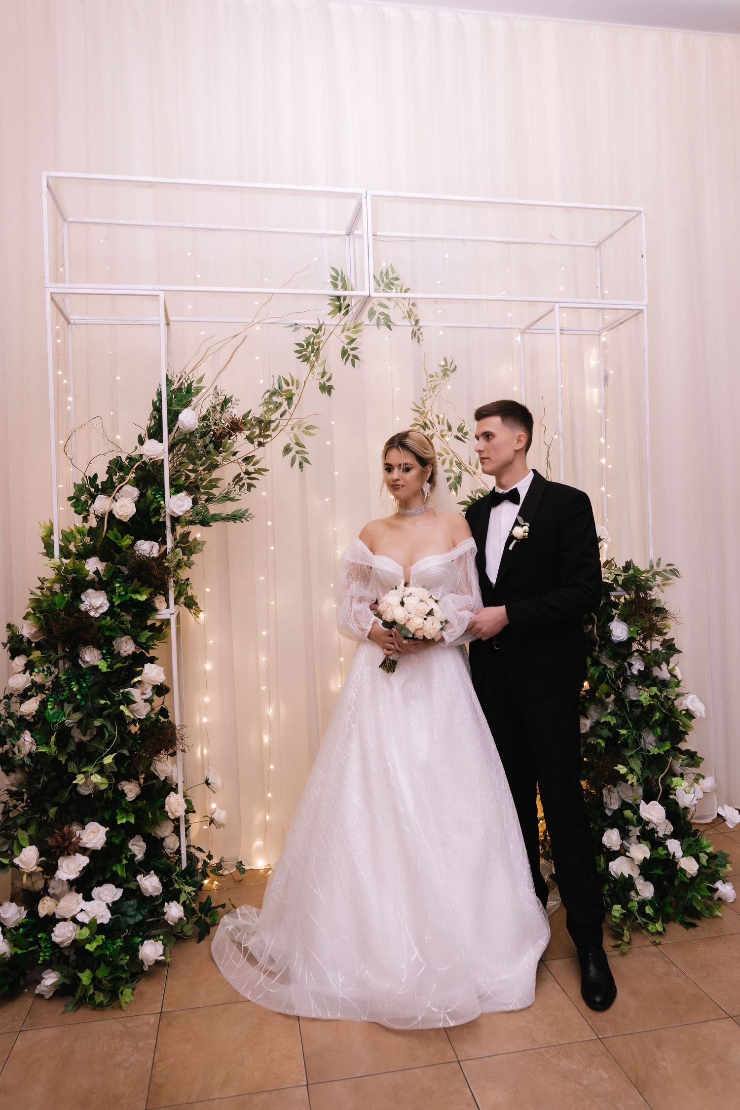 A couple poses in front of a floral arch with twinkling lights during their wedding celebration.