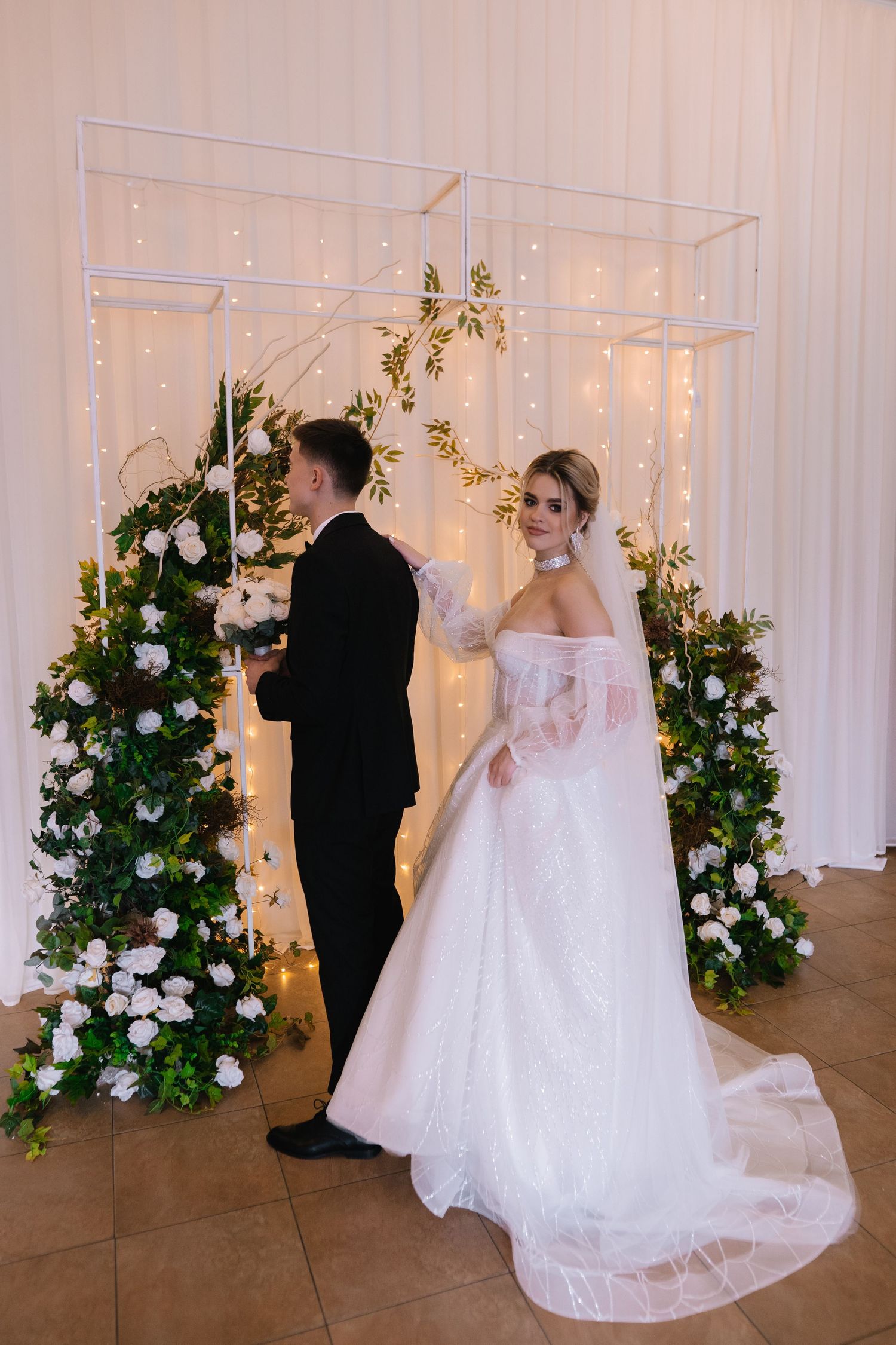 Couple at wedding ceremony standing by modern acrylic arch decorated with white flowers and twinkle lights.