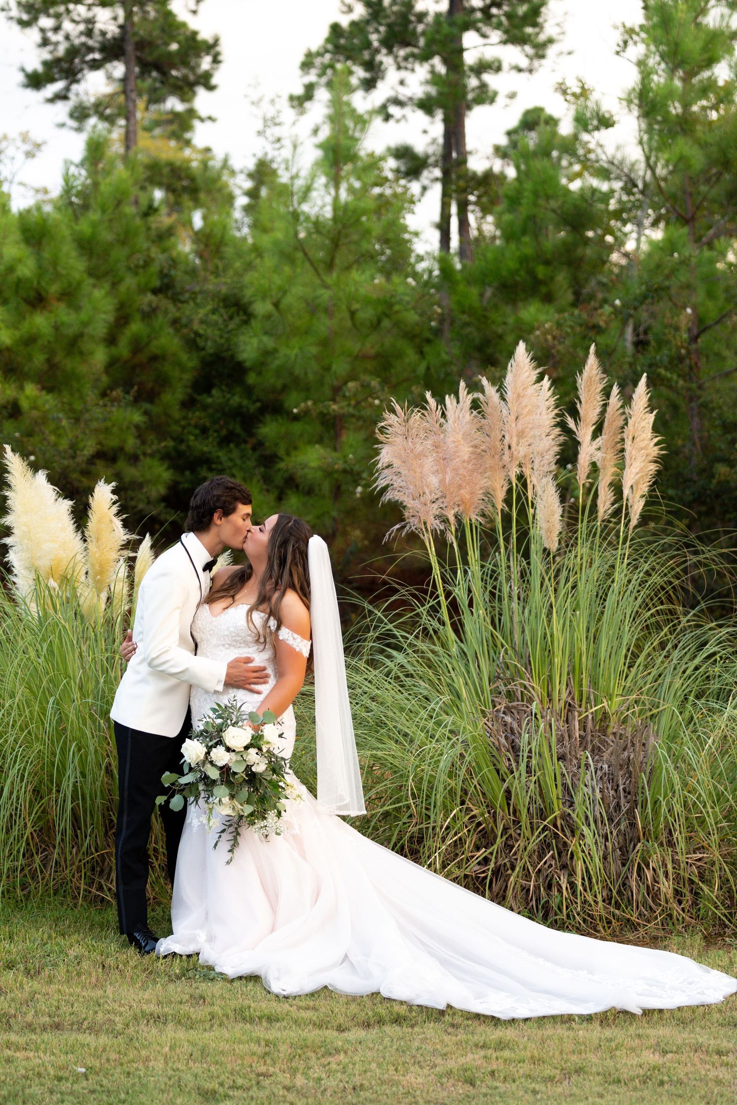 Romantic outdoor wedding scene with couple embracing near tall ornamental grasses and pine trees in natural setting.