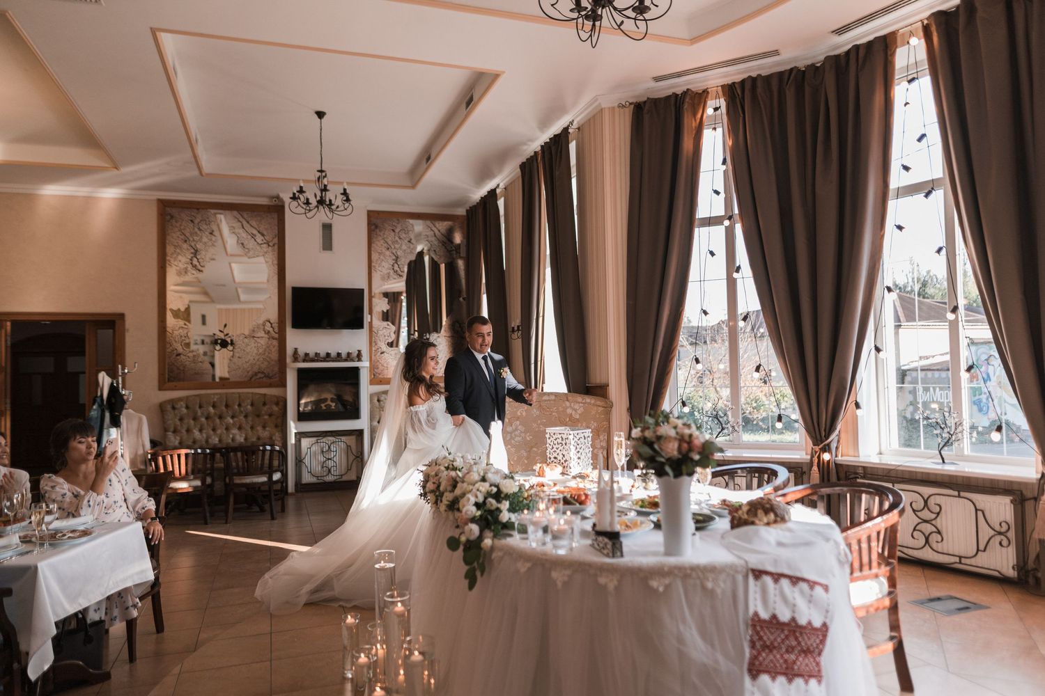 Elegant wedding reception setup in a bright room with tall windows, draped curtains, and decorated dining tables.