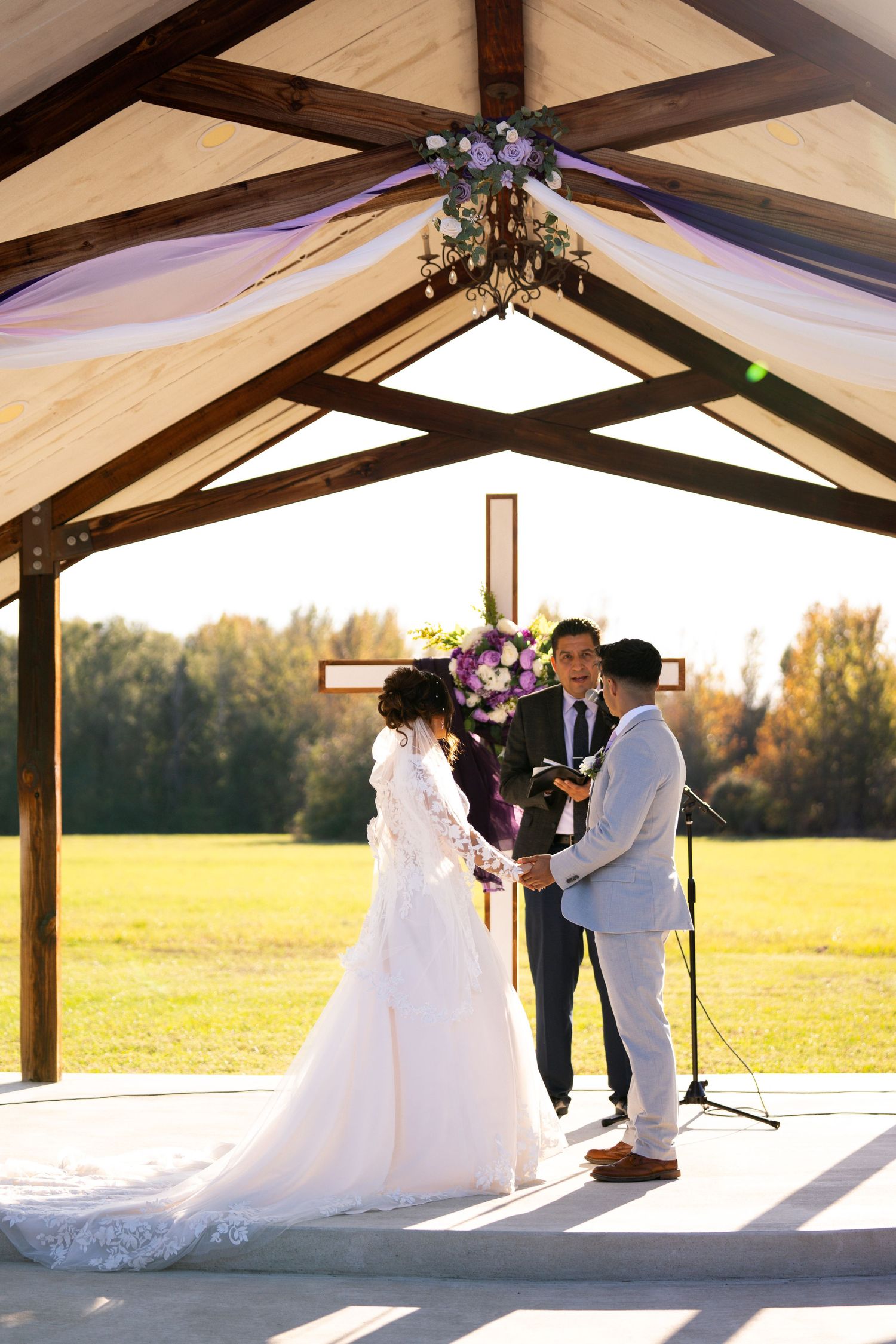 A wedding ceremony takes place under a rustic wooden pavilion with exposed beams during golden hour.
