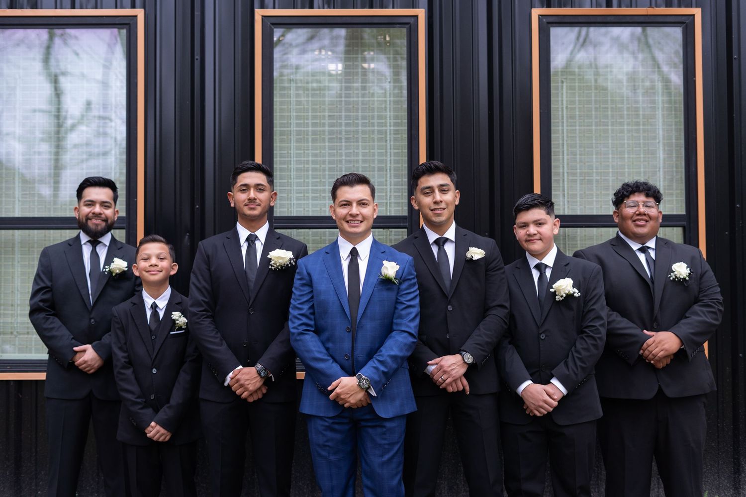 A wedding party in formal attire stands in a line against a building exterior with framed windows.