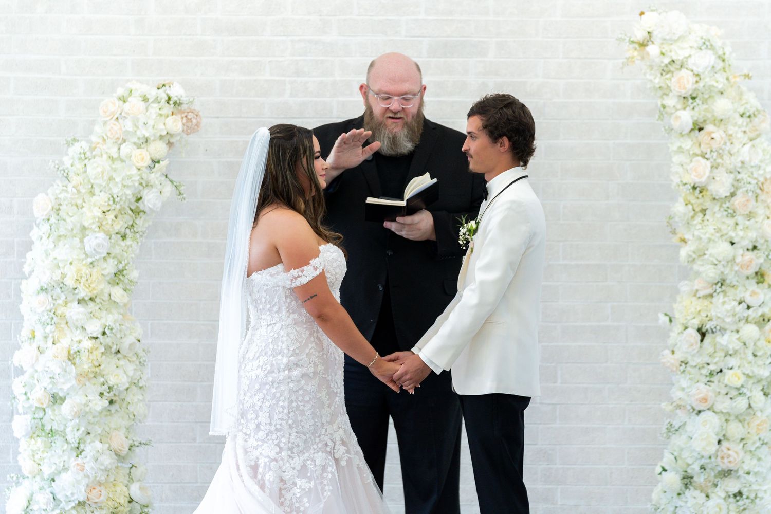 A wedding ceremony moment captured in front of an elegant white floral arch backdrop.