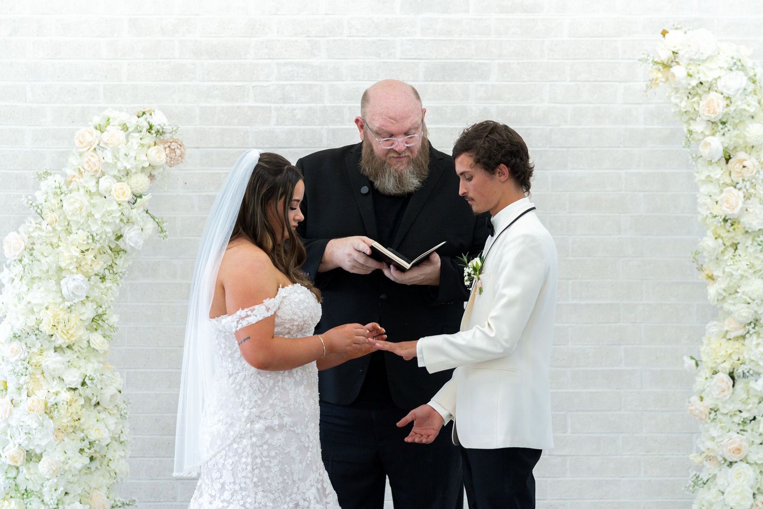 A couple exchanges vows during an outdoor wedding ceremony with white floral decorations in the background.