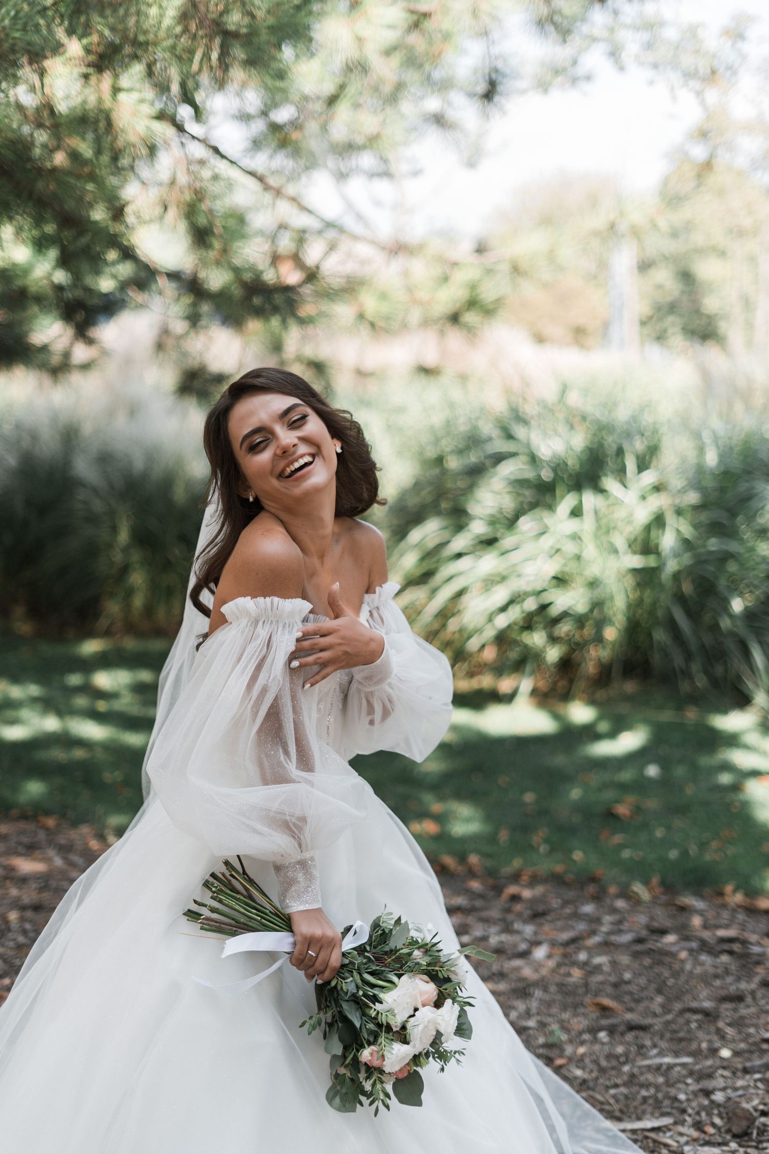 A joyful bride in an off-shoulder wedding gown holds a white and green bouquet in a garden setting.