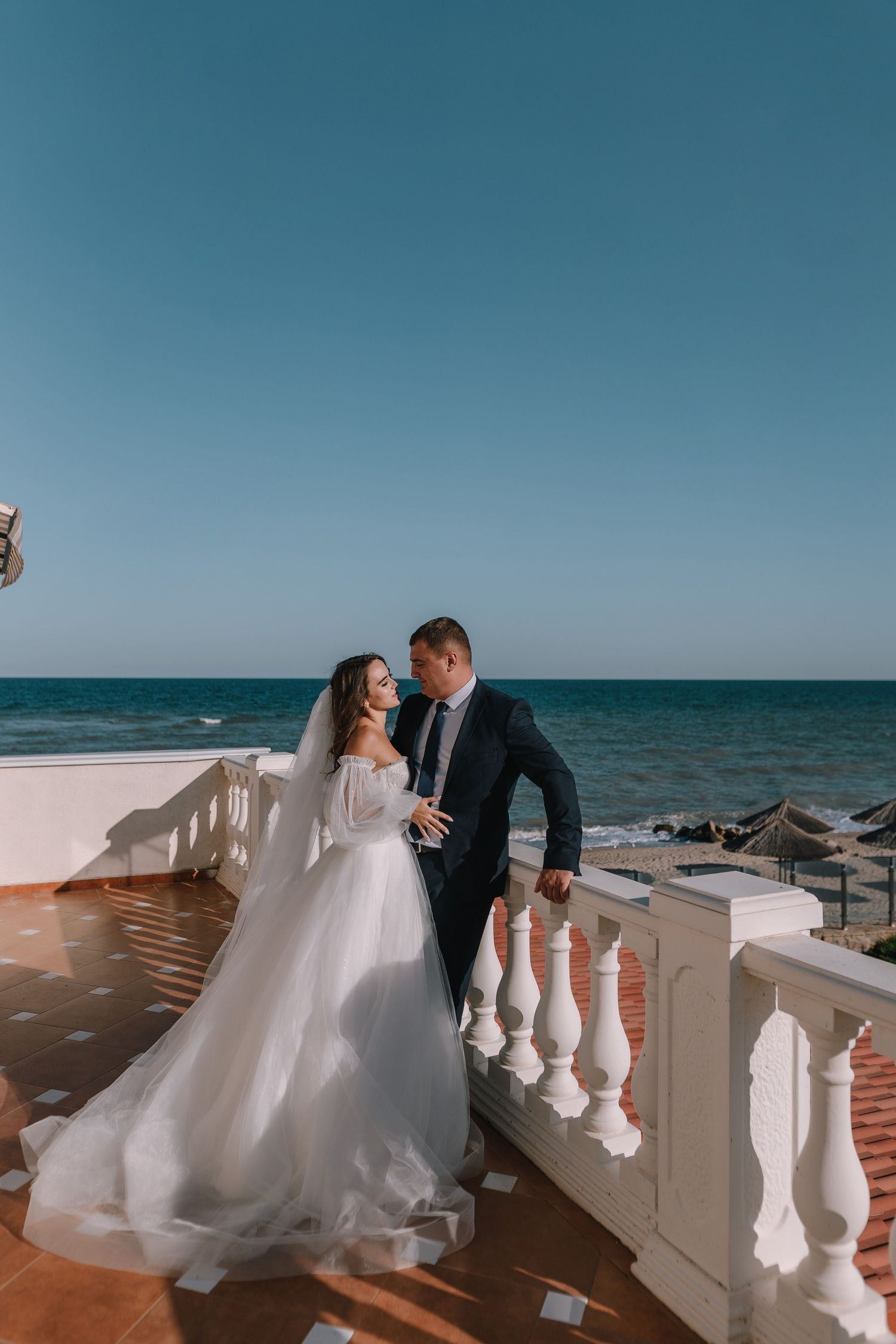 A romantic seaside wedding photoshoot featuring a couple posing along a white balustrade overlooking the ocean.
