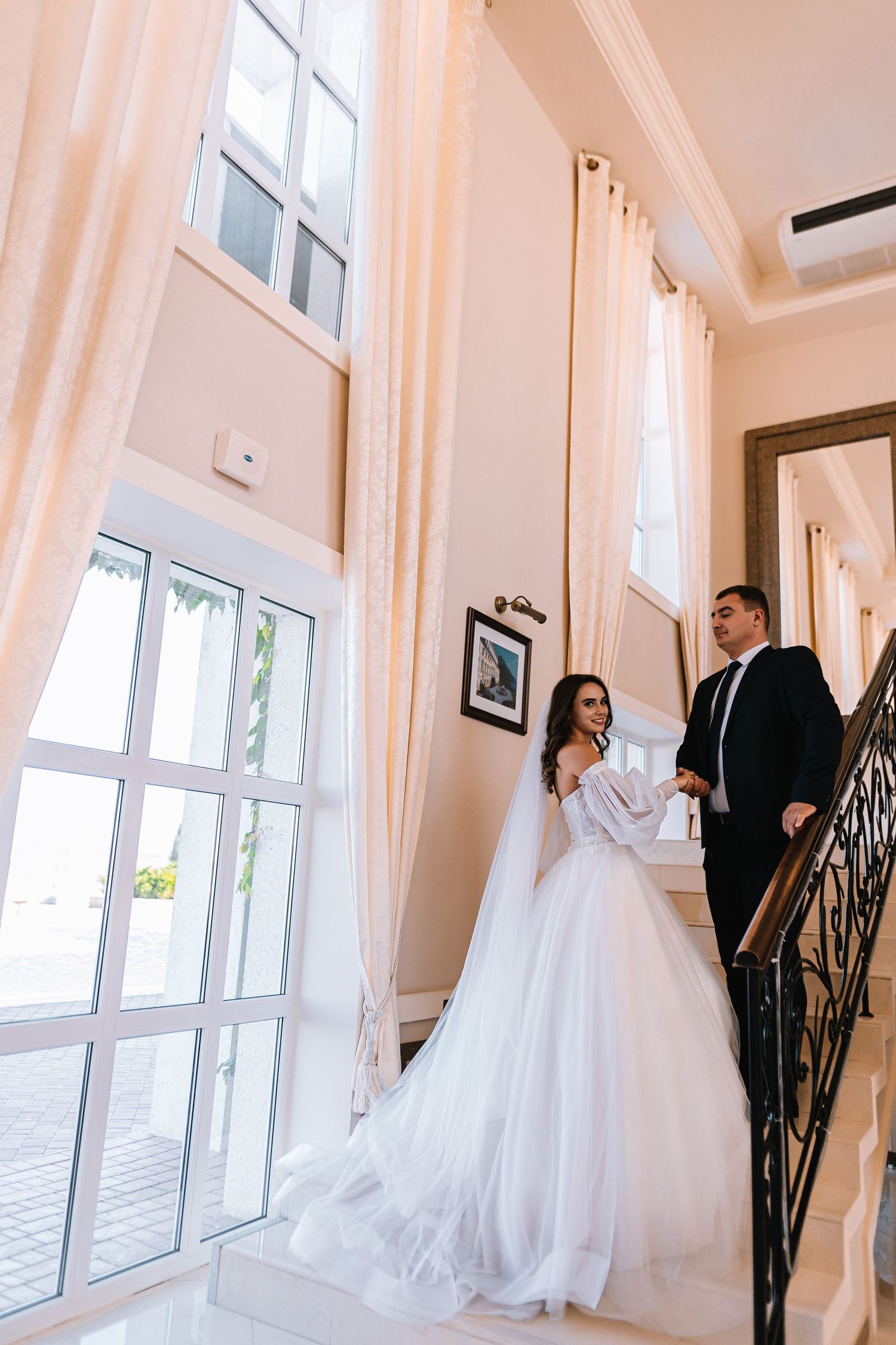 A couple in wedding attire stand on an elegant staircase with tall windows and white curtains in a bright interior.