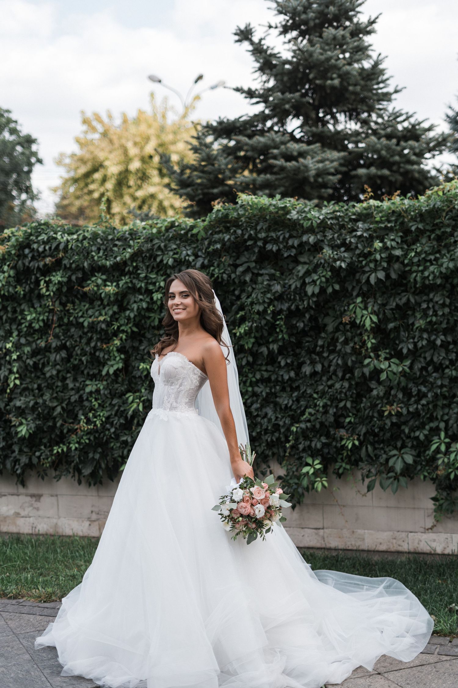 A bride in a flowing white ballgown poses gracefully against a backdrop of evergreen hedges and trees.