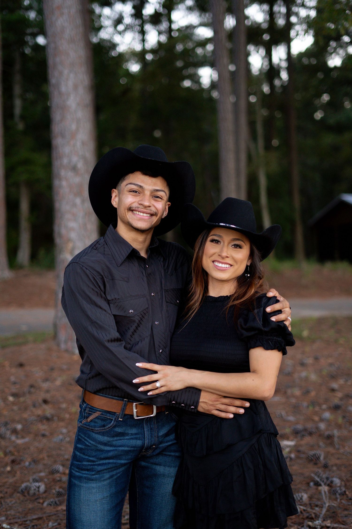 A couple in matching black outfits and cowboy hats pose together in a pine forest.