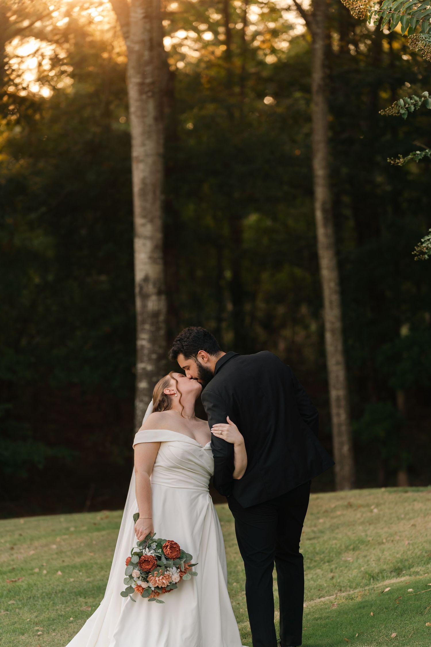 A bride in an off-shoulder gown and groom share an intimate moment at sunset among birch trees.