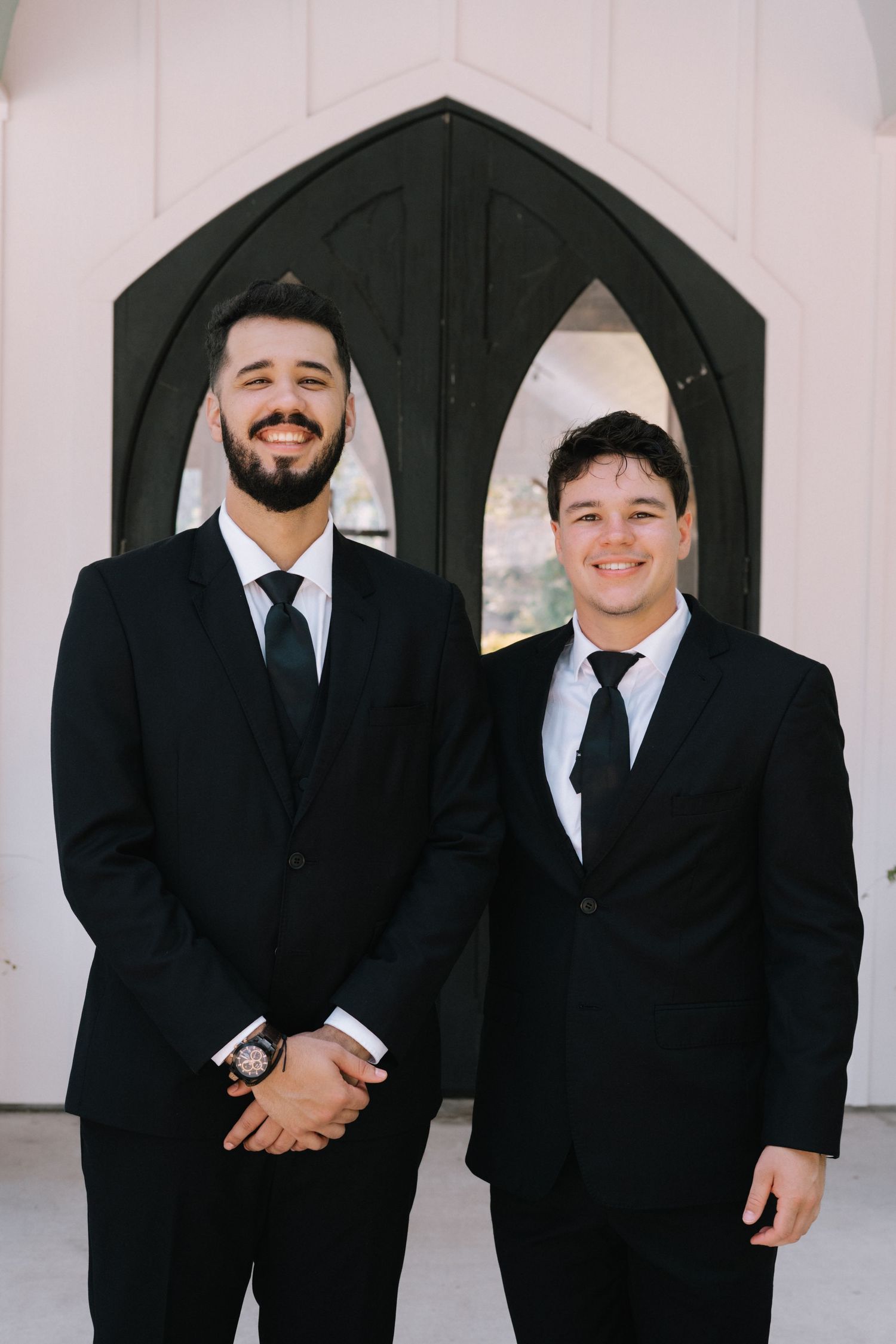 Two people in black suits and ties stand smiling in front of Gothic-style arched doorways.