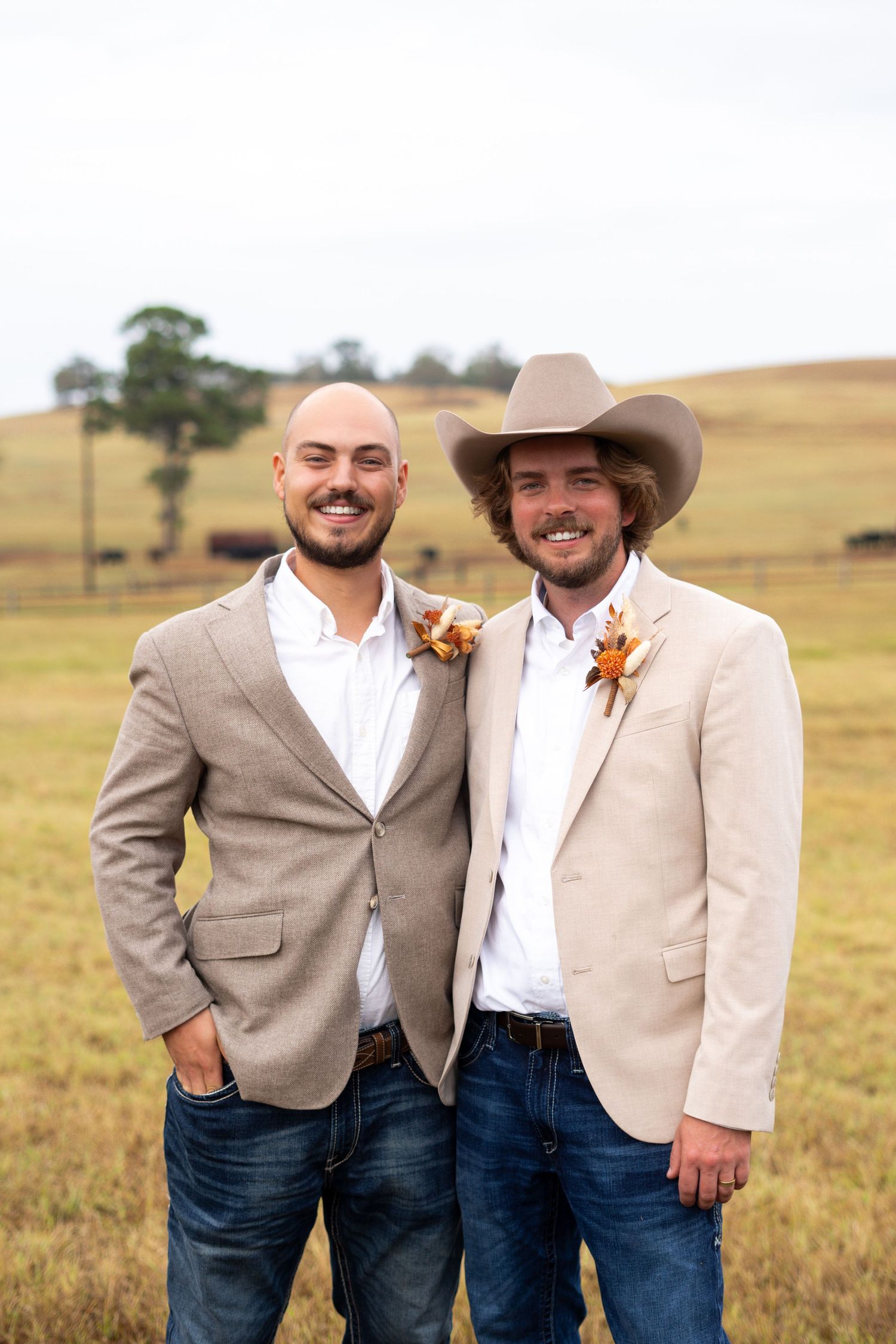 Two people in coordinating beige blazers and boutonnieres pose together in a pastoral field setting.